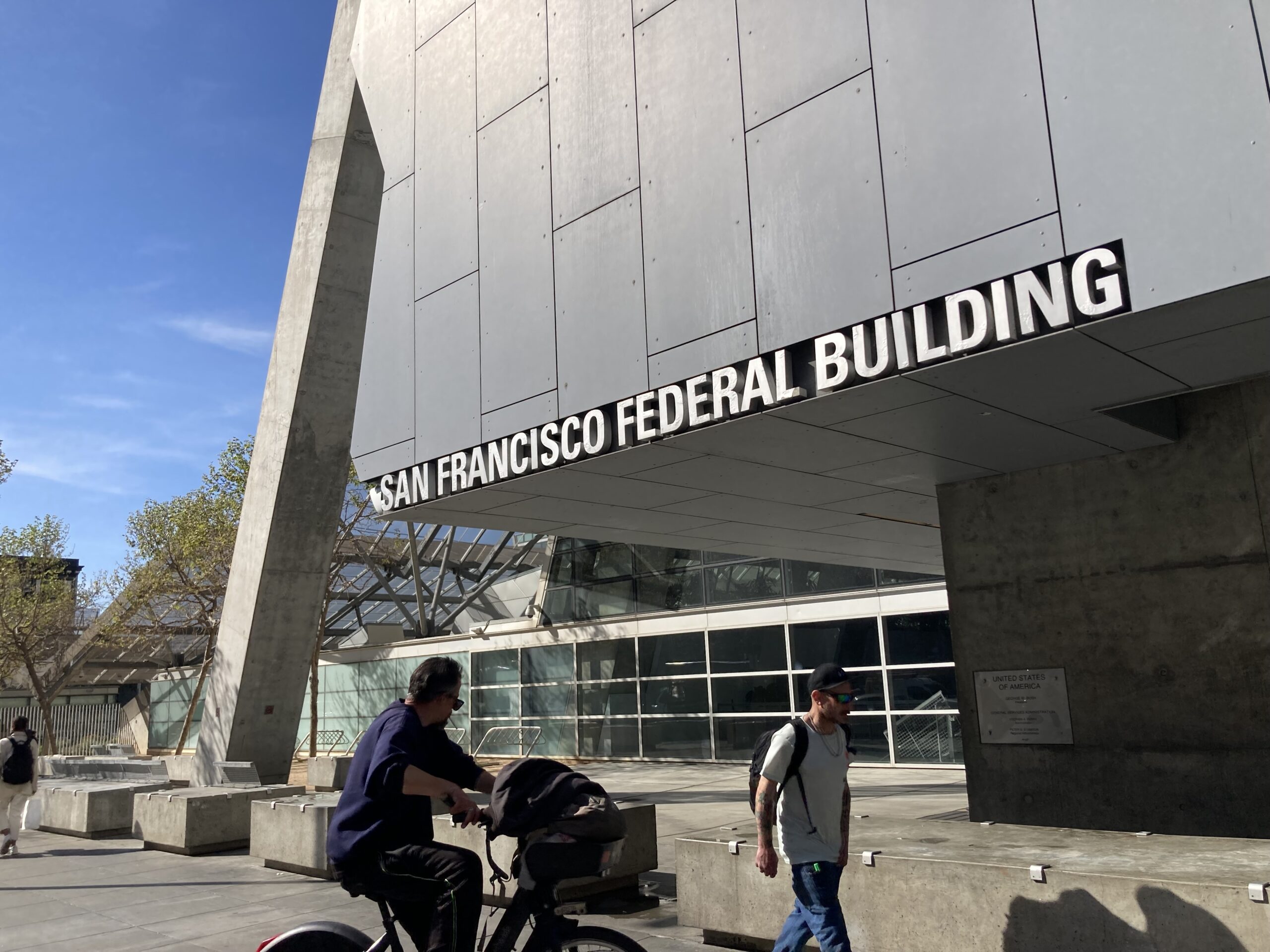 The image shows the San Francisco Federal Building with modern architecture. A cyclist and a pedestrian are passing by under a clear blue sky.