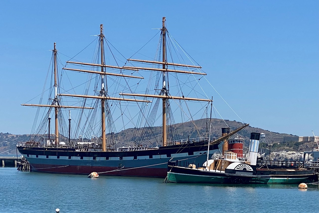 A historic looking long sailboat is docked next to a classic riverboat with Alcatraz island in the background.