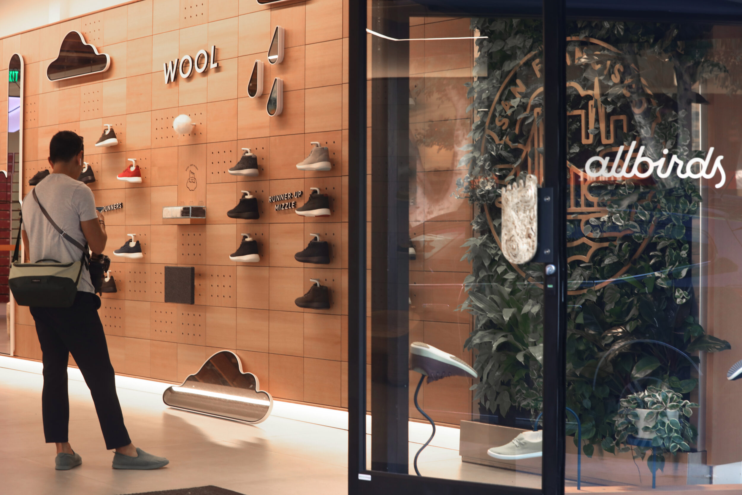 A person examines a shoe display inside an Allbirds store with a wood-inspired decor and greenery.