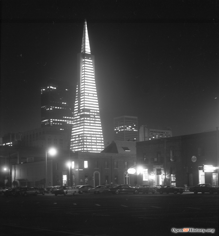 A black-and-white night shot of a lit skyscraper towering over surrounding buildings.