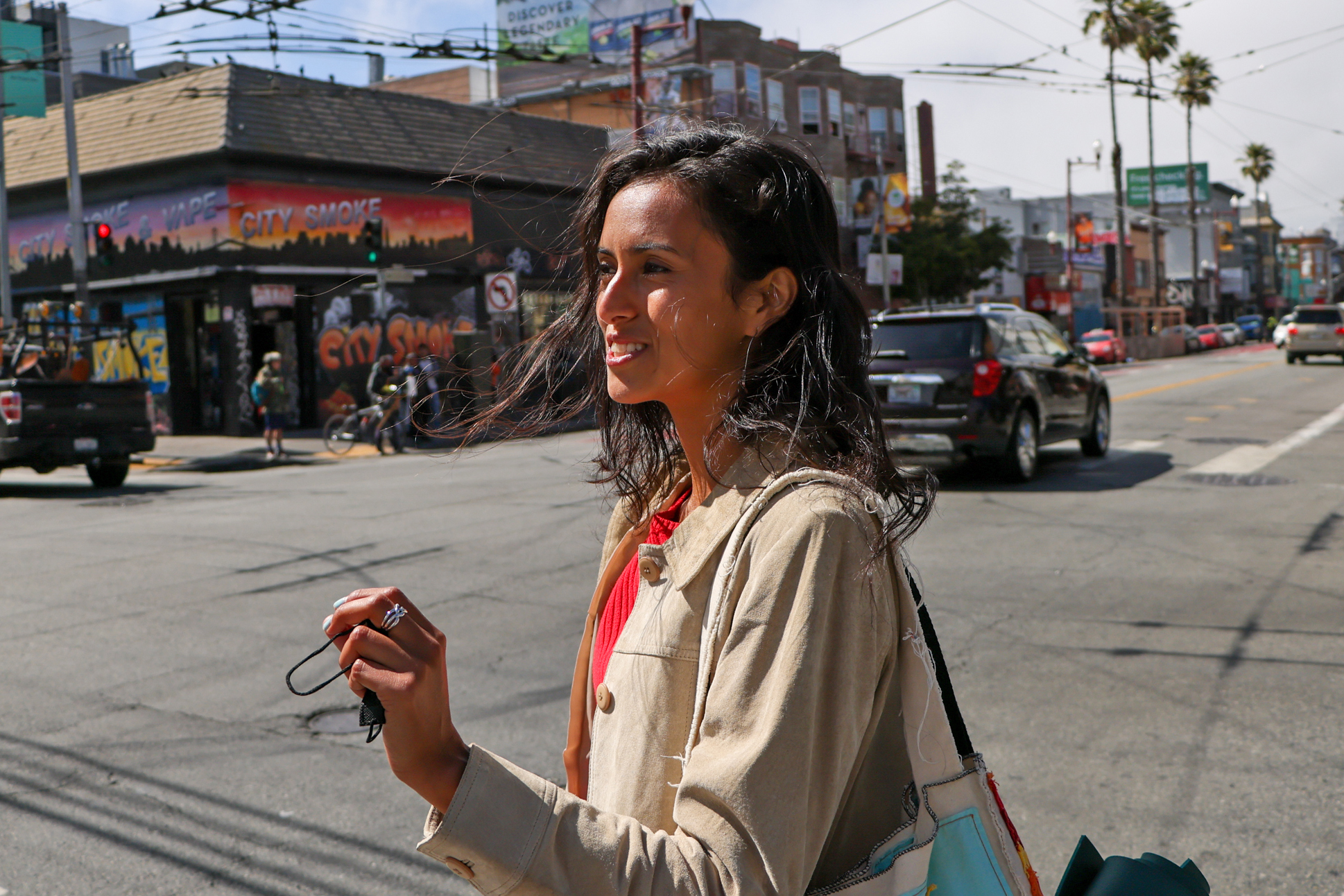 A person stands at a busy city intersection, holding something in their hand. Behind them, colorful storefronts and street art are visible, along with cars and palm trees.