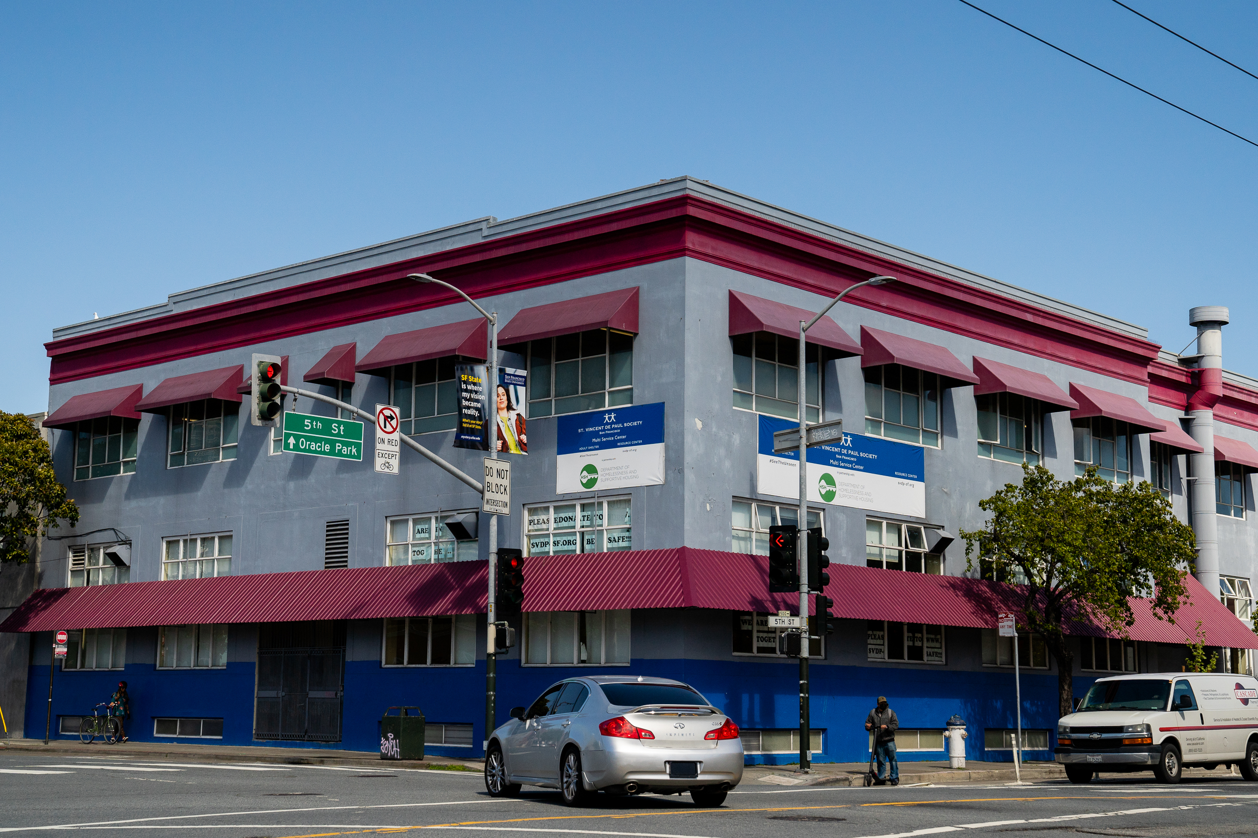 The image shows a large, corner building with gray and blue walls and a red roof. There are several windows with red awnings, a few trees, and traffic lights.