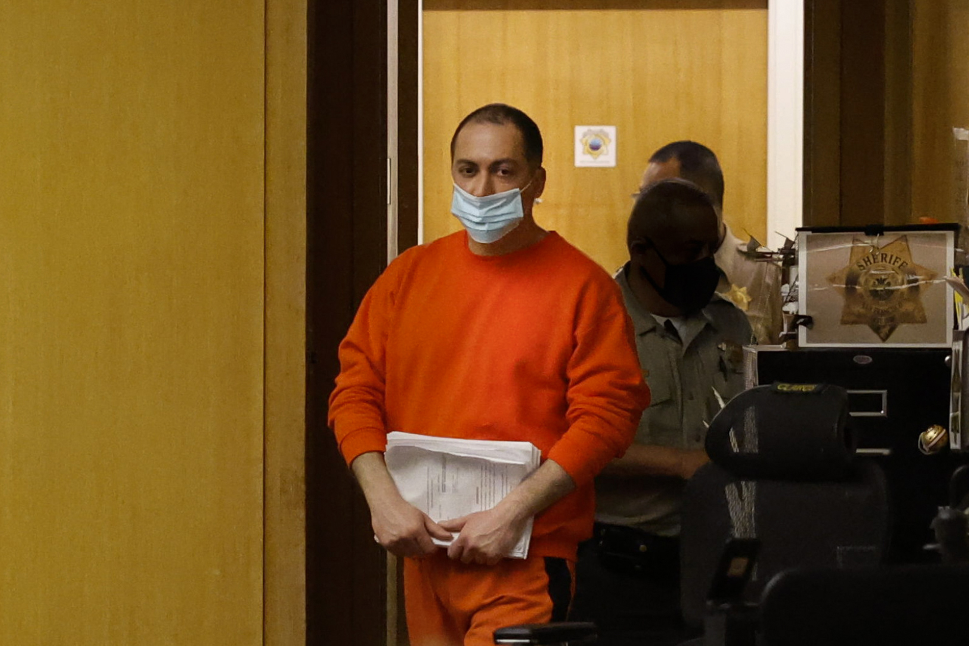 A man in an orange jumpsuit and a light blue face mask is holding papers, walking with a security officer in uniform. They are in a room with wooden walls.