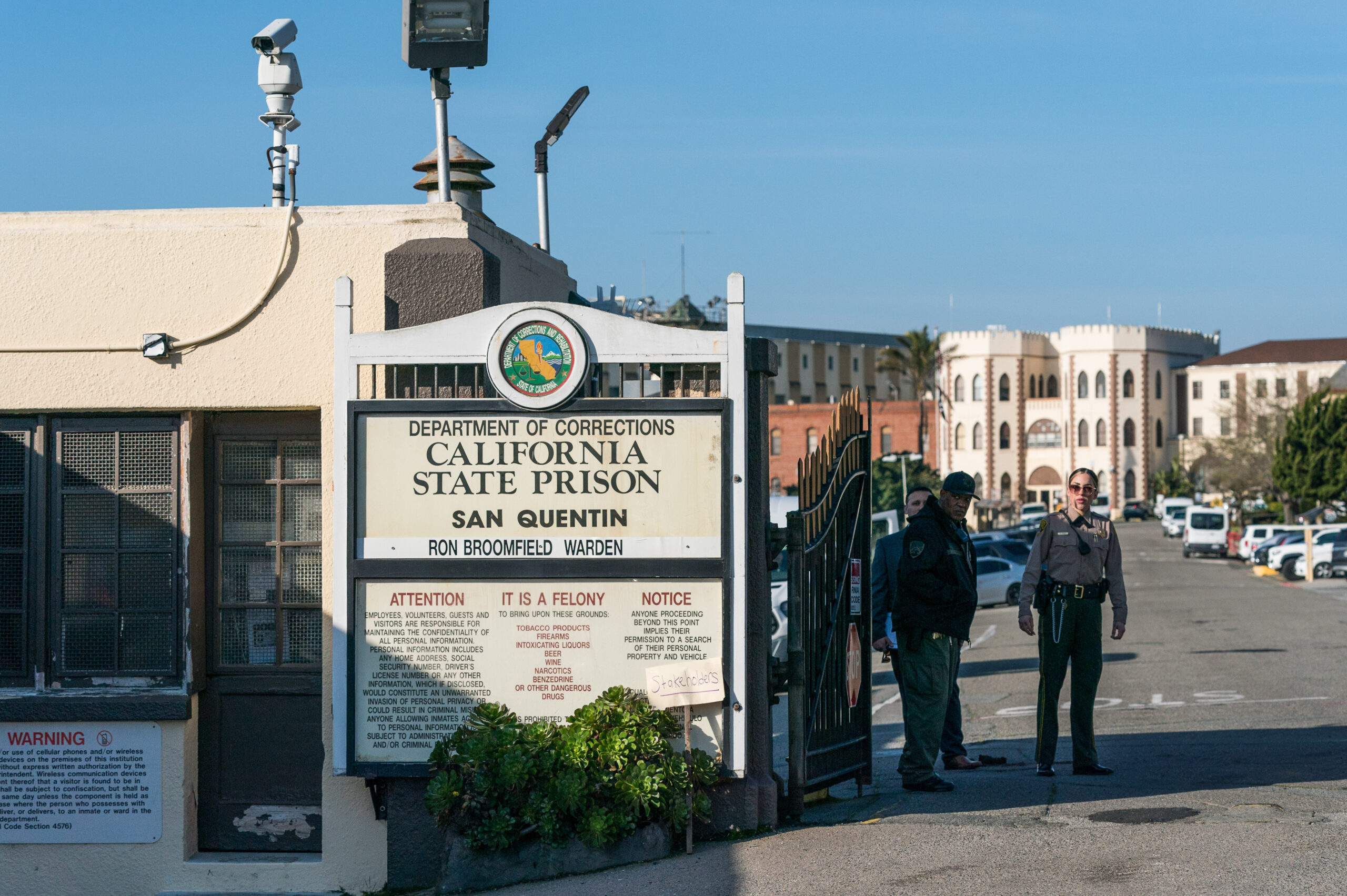 Two officers stand by the entrance of San Quentin State Prison, with a sign and security cameras nearby.
