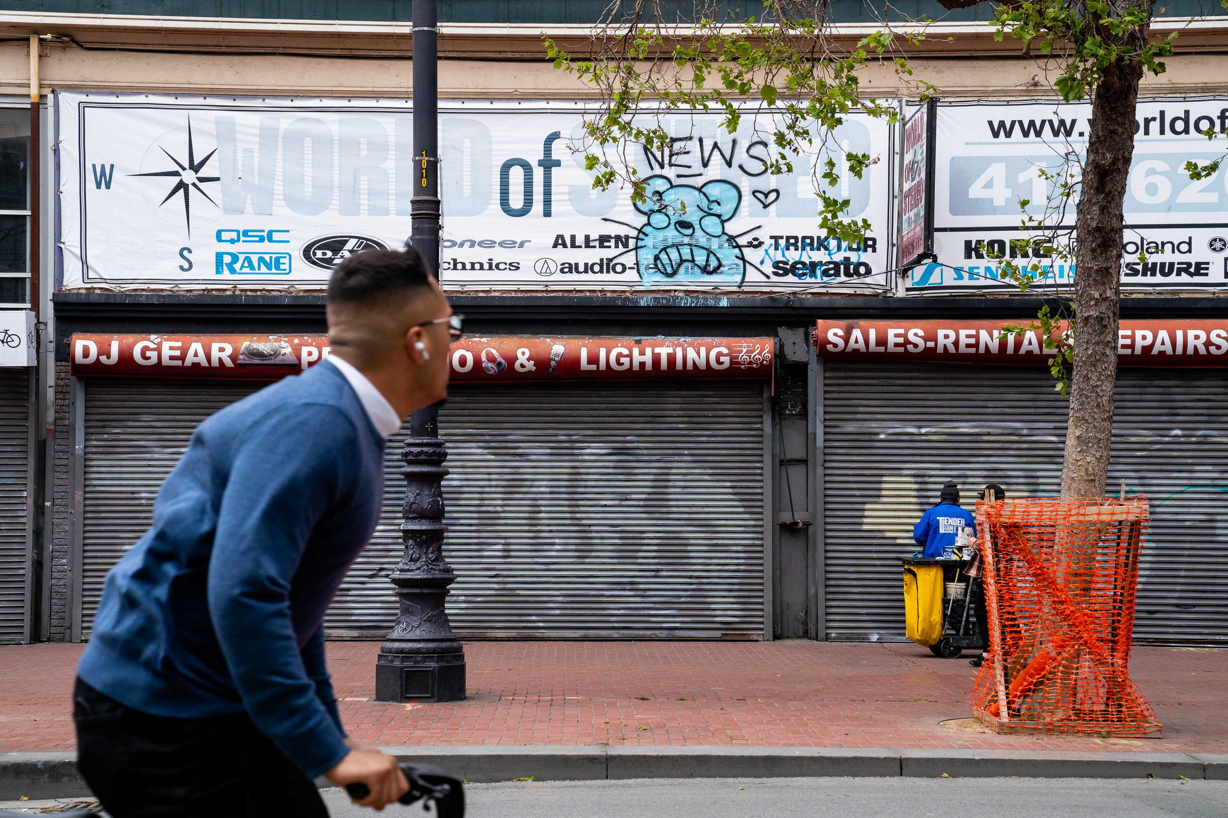 A person on a bike wearing earbuds rides past a closed store with &quot;DJ Gear &amp; Lighting&quot; signage. In the background, a worker pushes a cart near an orange barrier.