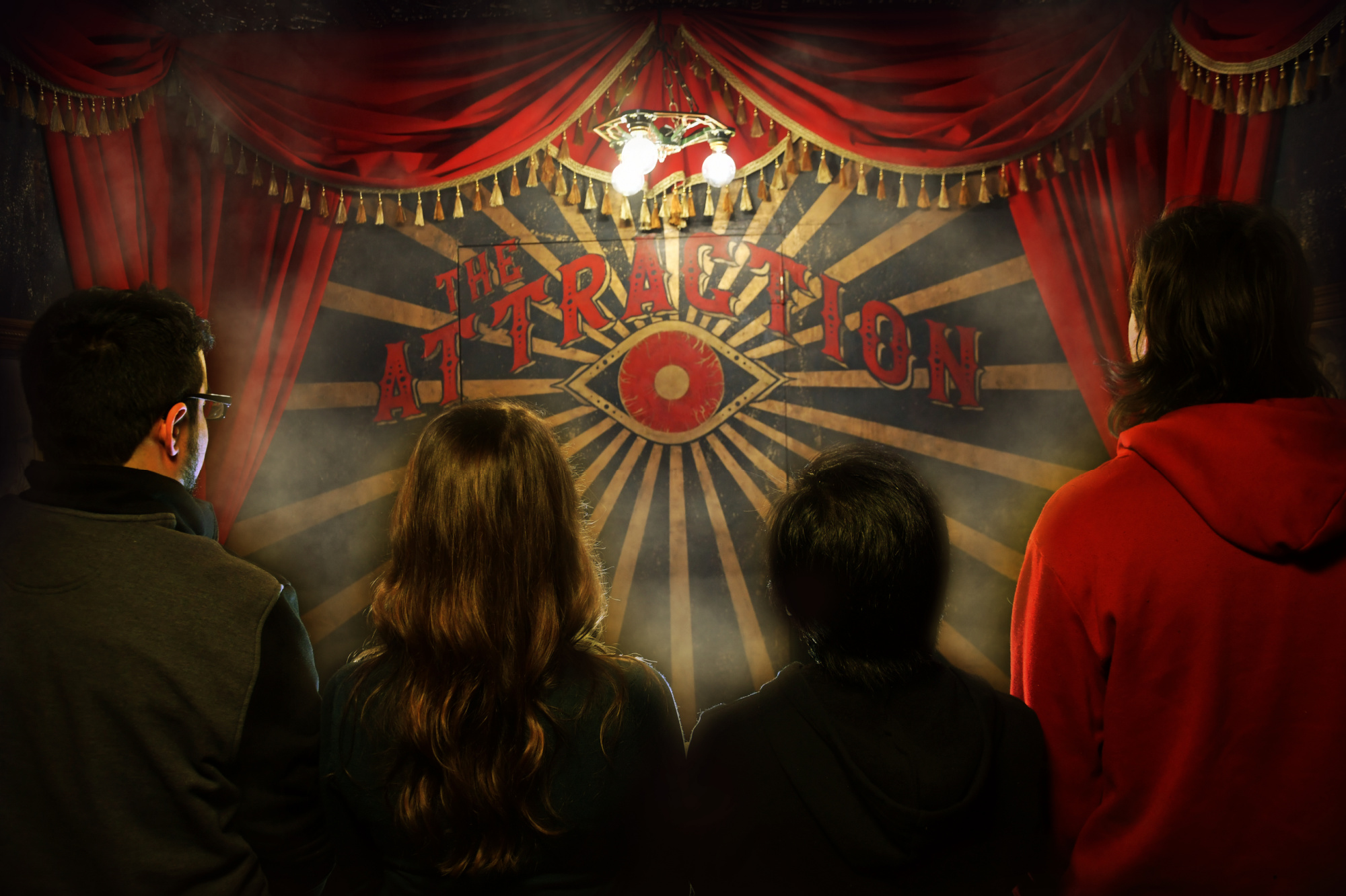 Four people stand facing the other way looking at an old-fashioned magic sign reading The Attraction above a red eyeball.