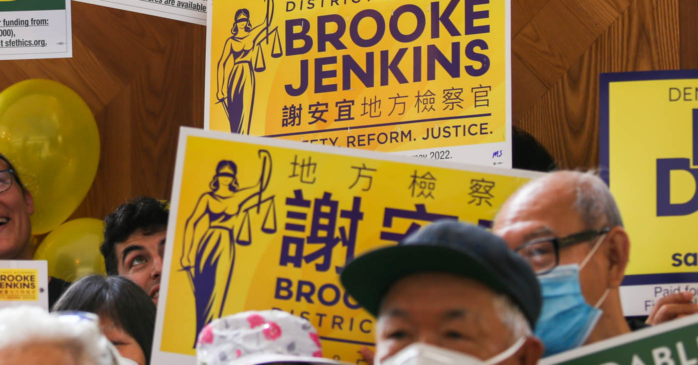 Supporters hold campaign signs during a rally for DA Brooke Jenkins that have text in both English and Chinese.