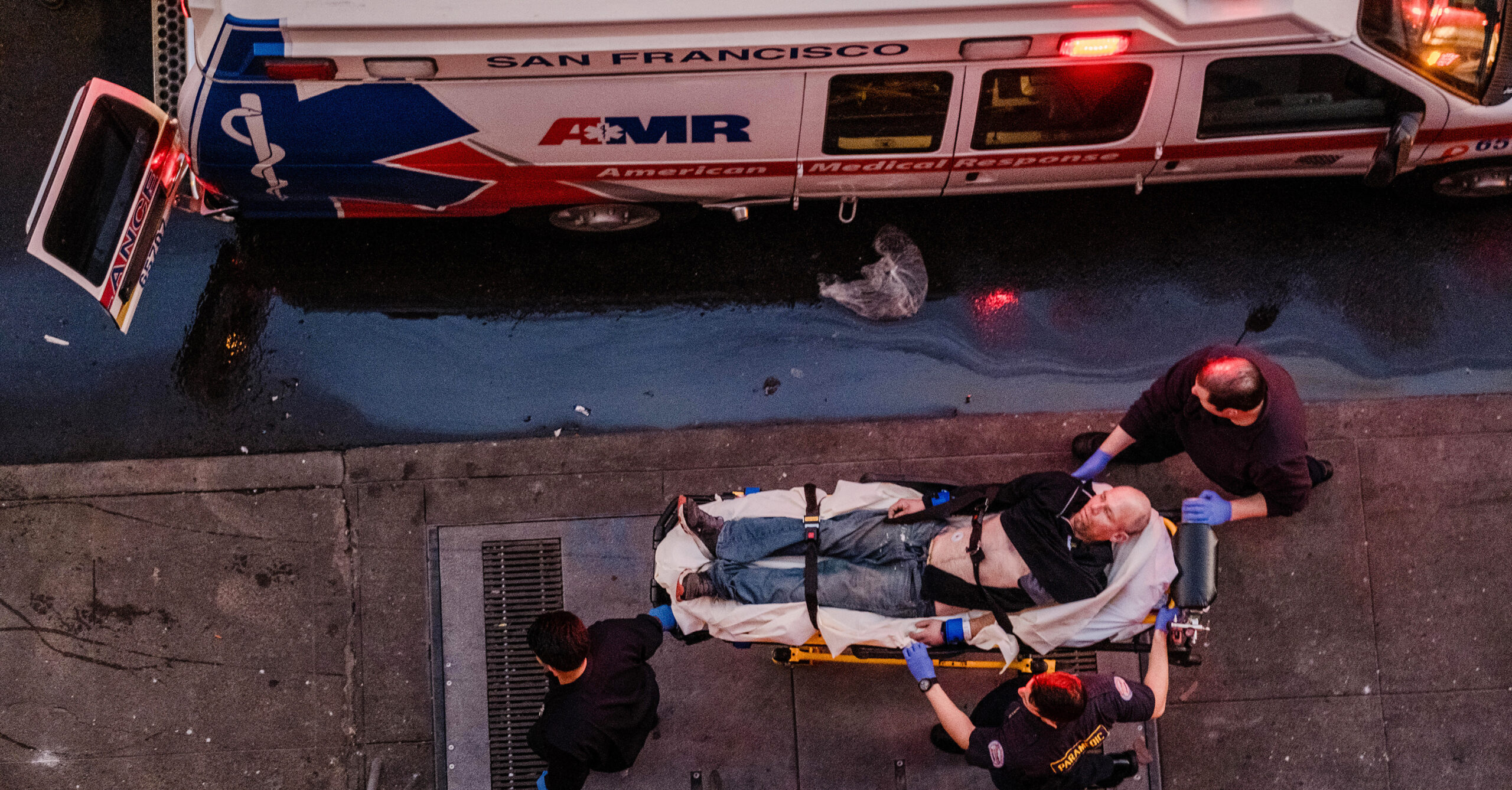 Paramedics attend to a person on a stretcher beside an ambulance on a city street at dusk.