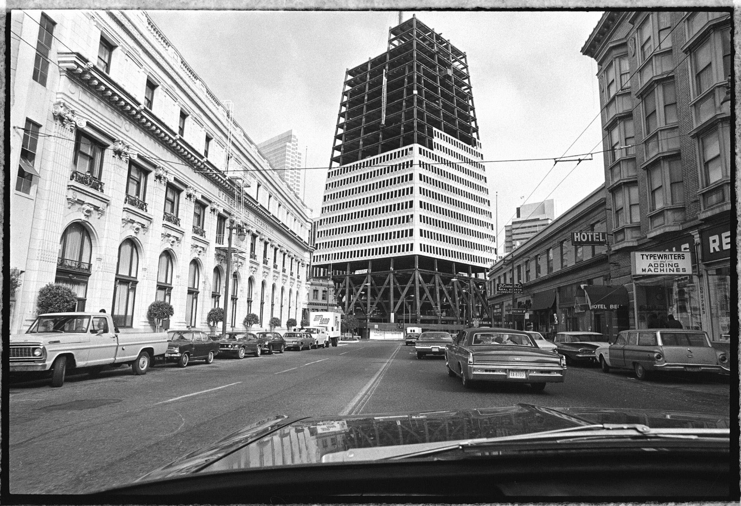 A vintage black and white photo of a busy street with old cars, classic buildings, and a half-finished skyscraper.