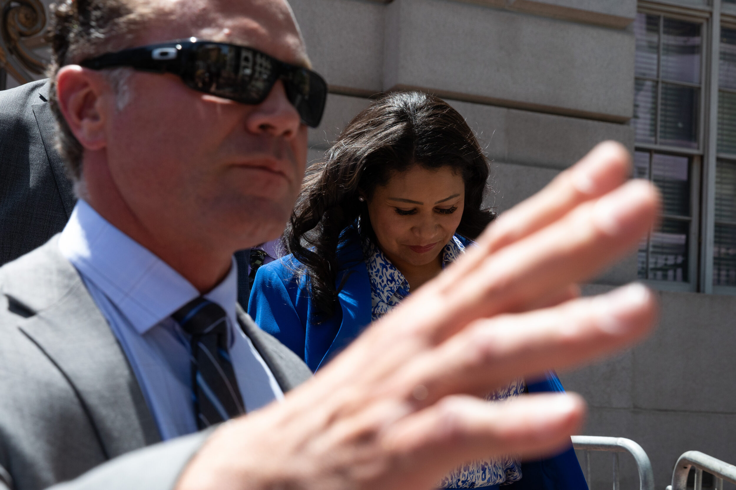 A man in sunglasses extends his hand to the camera; a woman in blue looks down behind him. They're outside, in daylight.
