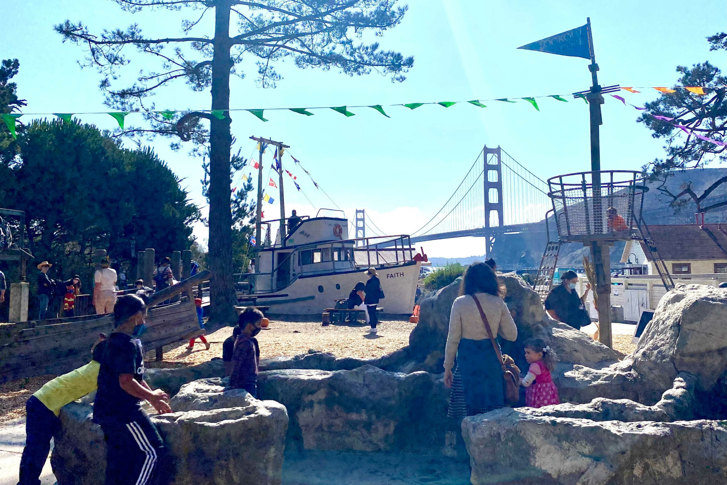 People milling around an outdoor kids' park with the Golden Gate Bridge in the background.