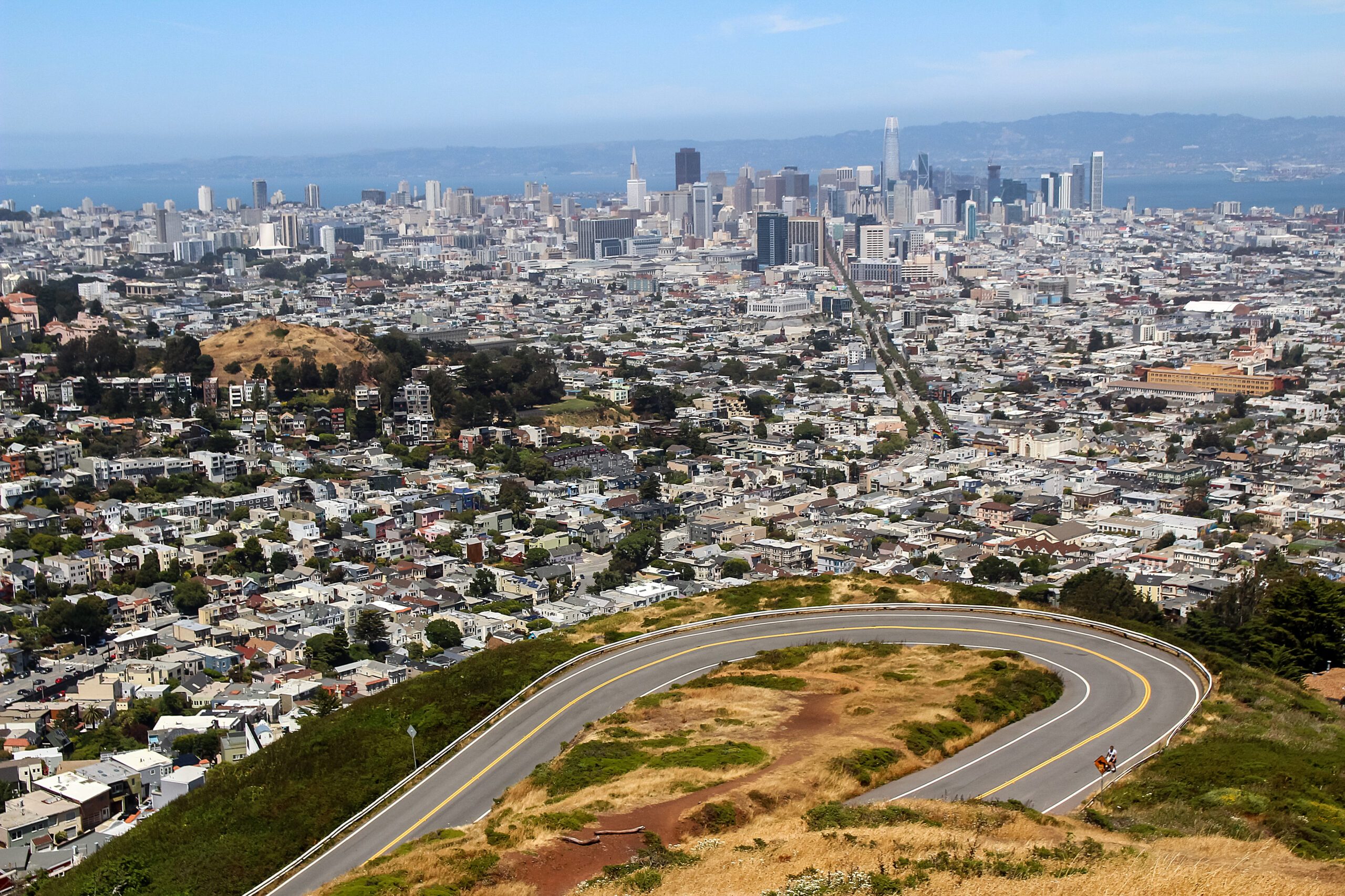 A cityscape with tall buildings in the distance, viewed from above a windy road on a hill, showing a mix of residential and commercial areas under a clear sky.