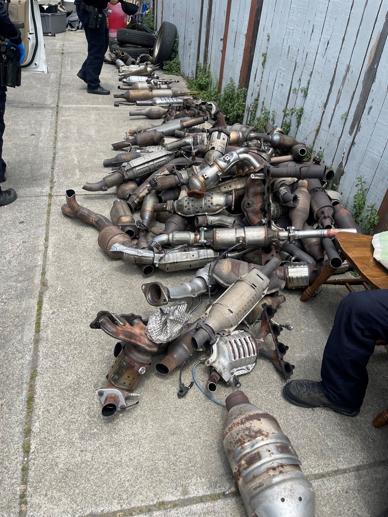 A large pile of catalytic converters found in raids are strewn across a sidewalk against a wooden fence.
