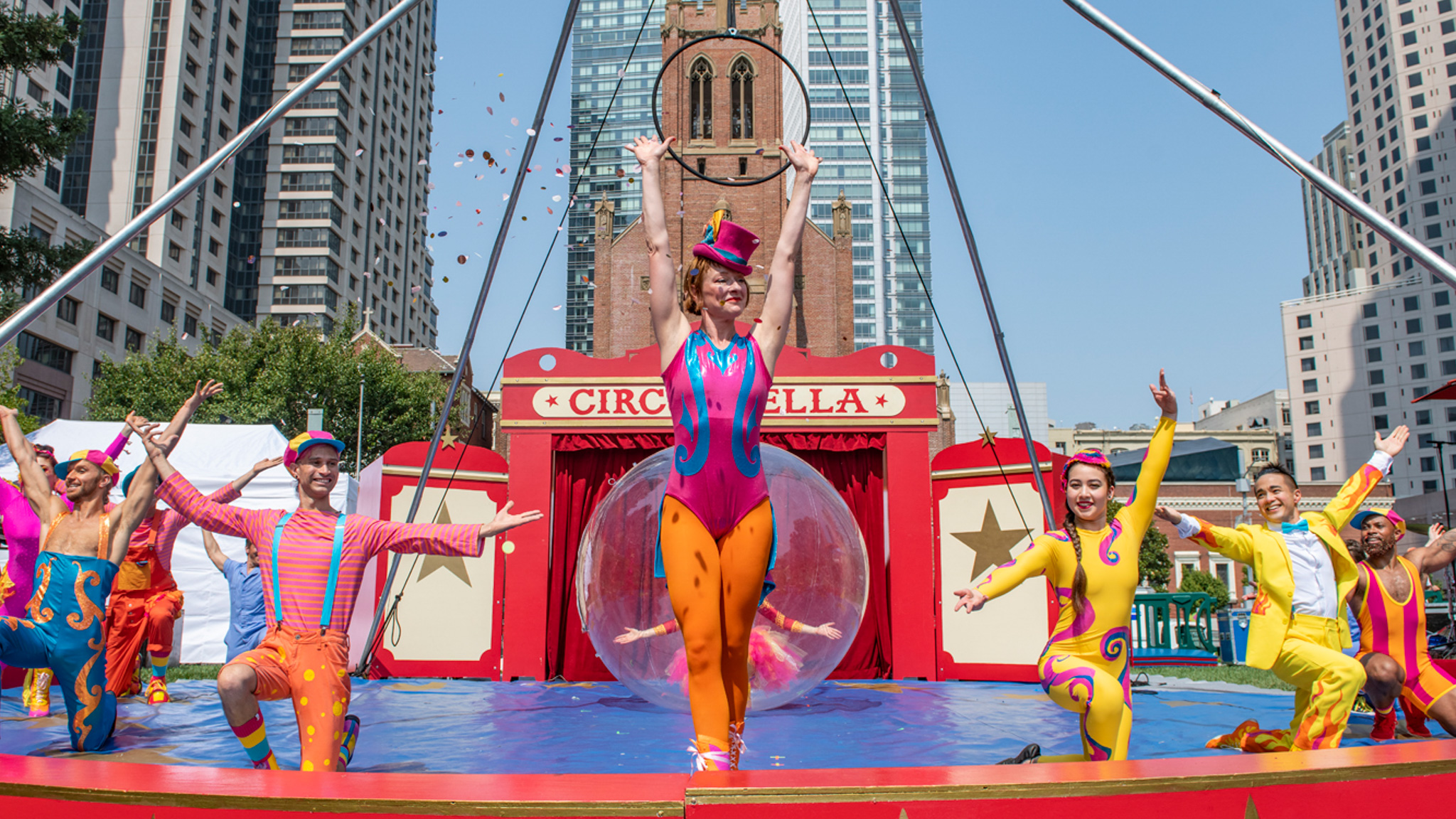 A trapeze artist stands on a red and blue stage with circus performers surrounding her. 