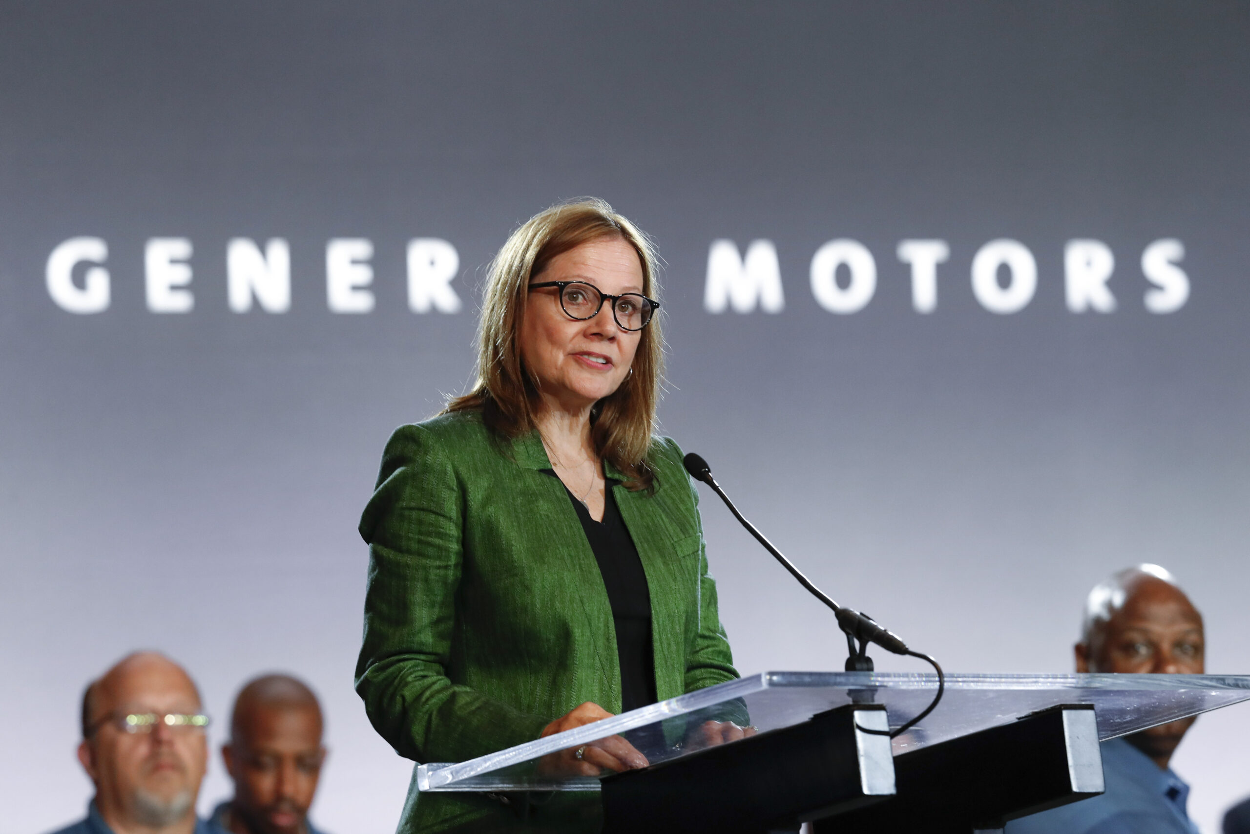 A woman in a green jacket at a podium in front of a general motors sign.