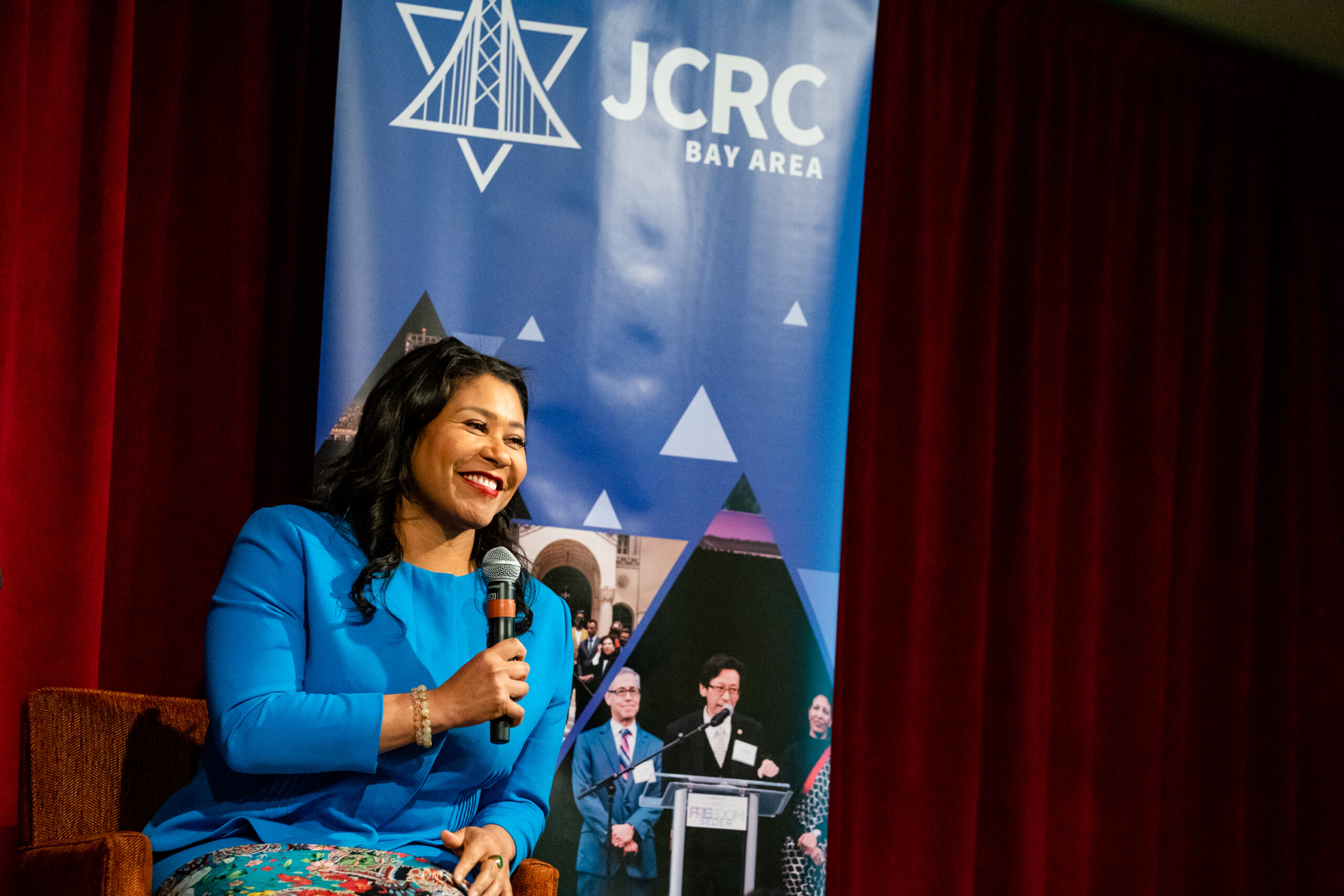 Mayor London Breed, wearing a blue dress, smiles and speaks into a microphone while seated by a banner of the JCRC Bay Area.