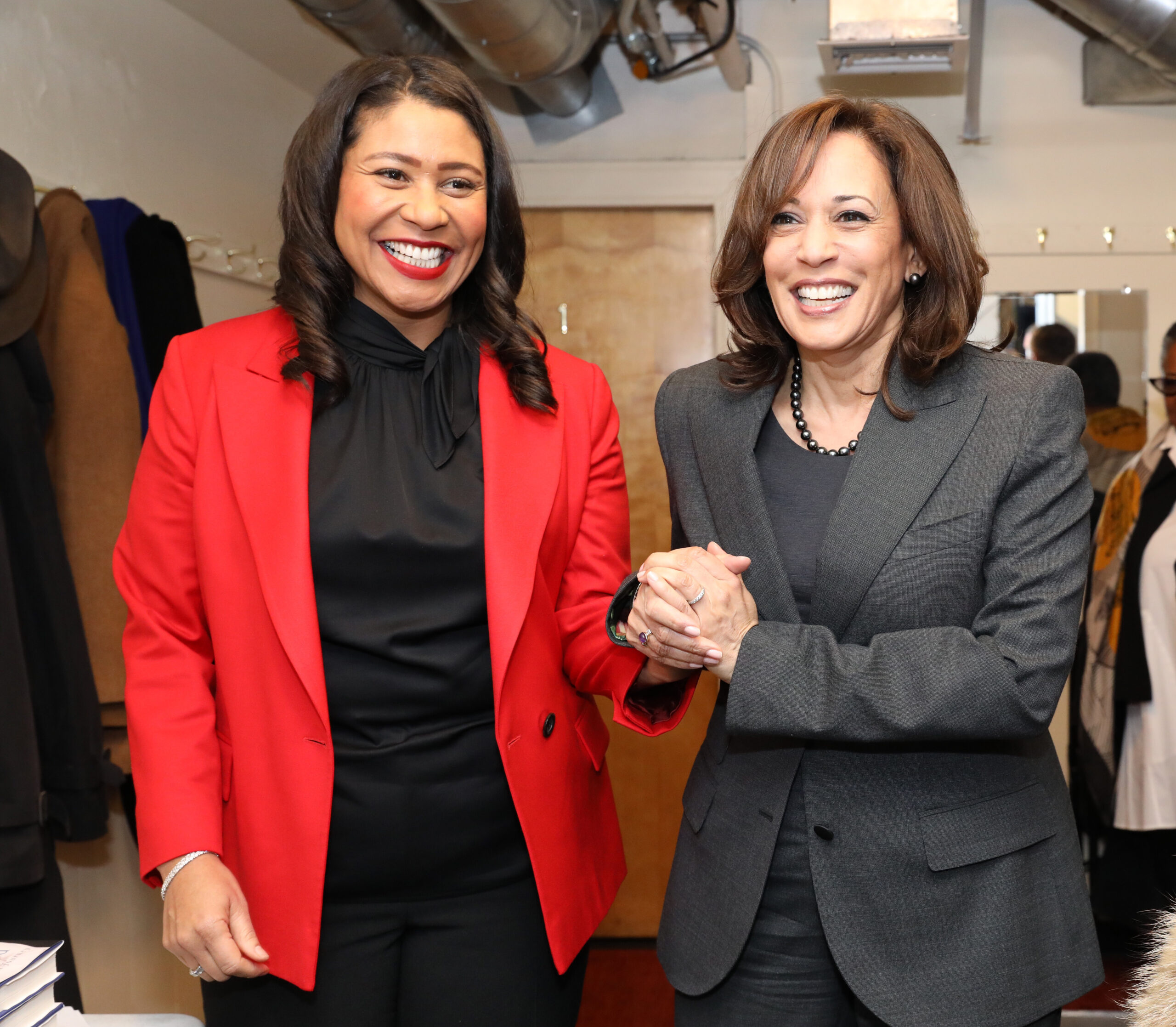 Two women are smiling and holding hands. One wears a red blazer with a black blouse and pants, and the other wears a dark gray suit with a black top and a pearl necklace.