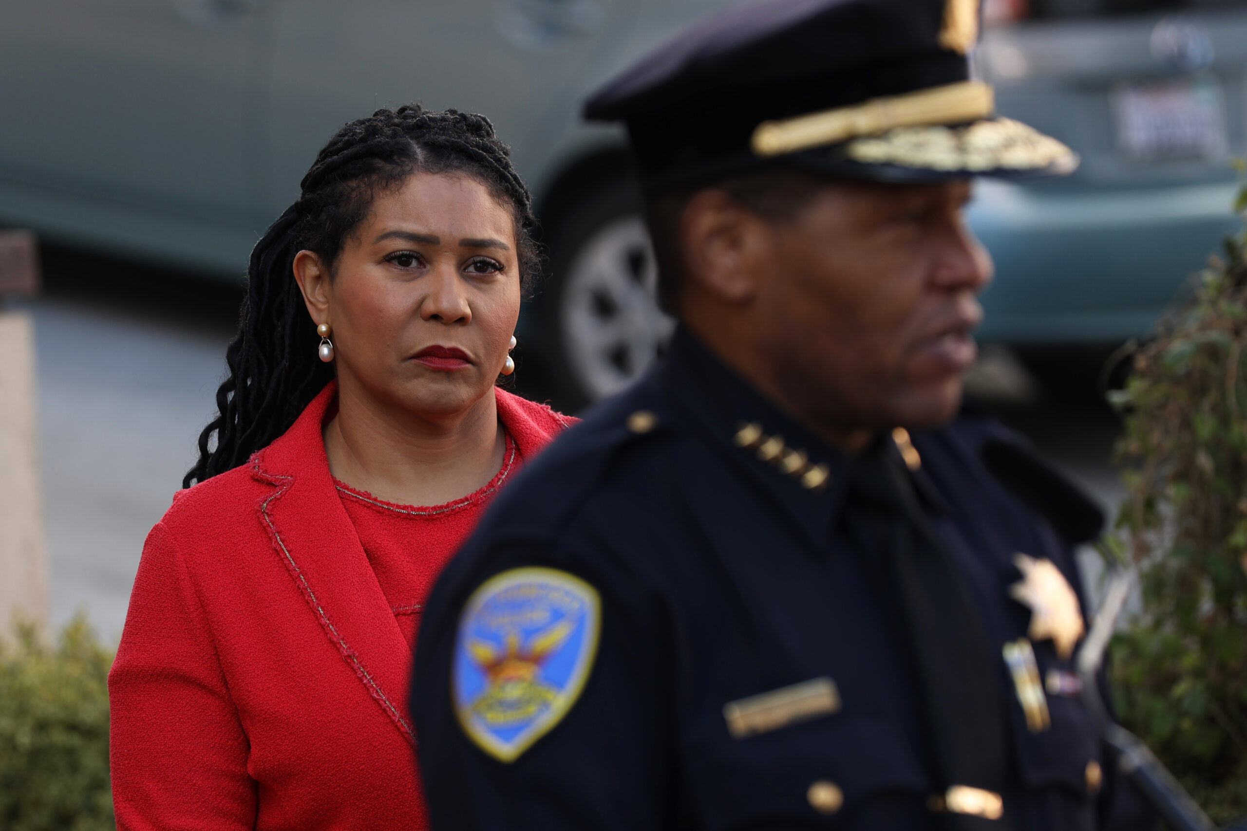 A woman in a red coat appears pensive beside a blurred man in police uniform.