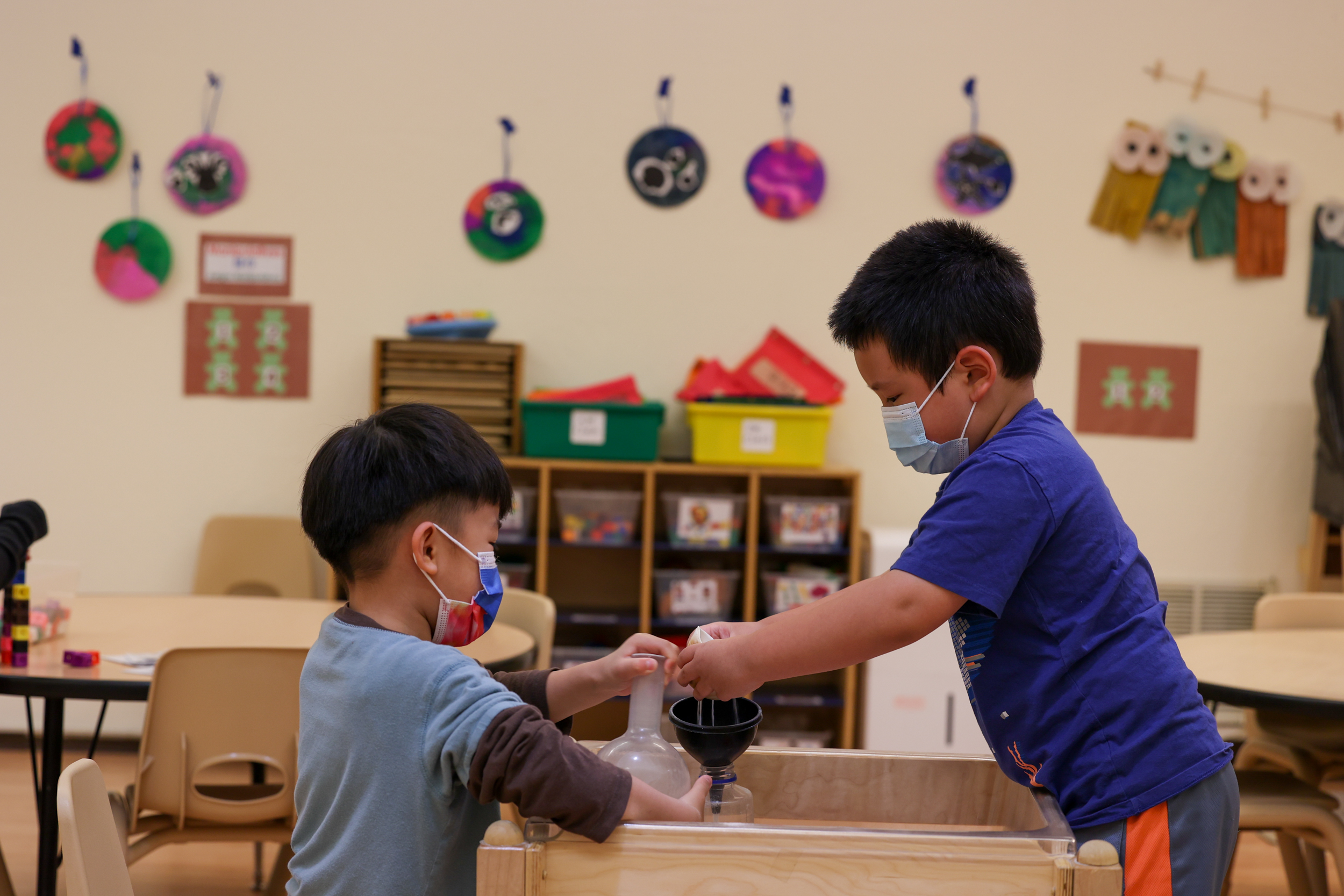 Two children wearing masks are playing with toys at a table in a classroom. Colorful artwork hangs on the wall behind them, and there are shelves with plastic bins.