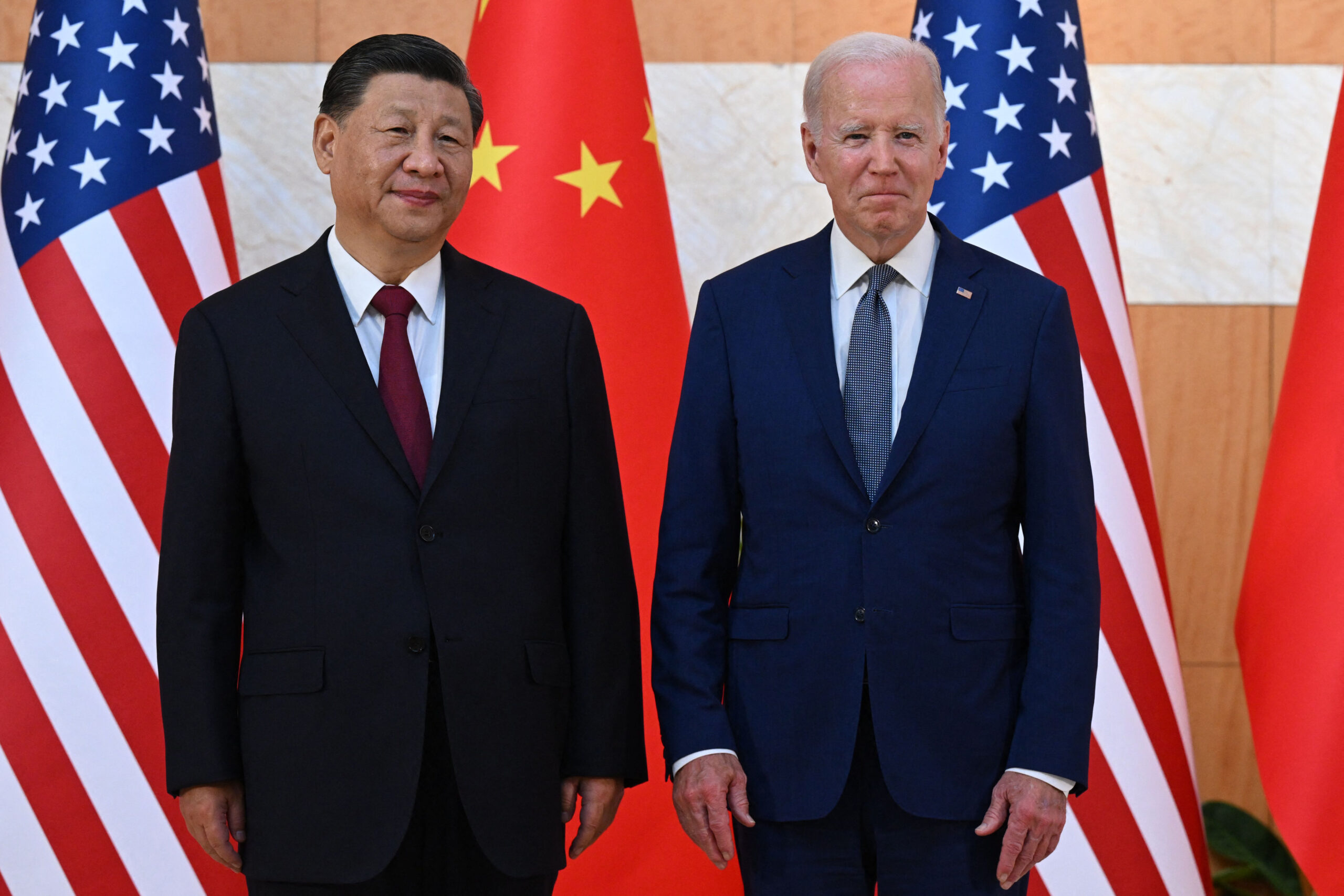 US President Joe Biden, right, and China's President Xi Jinping, left, wearing suits and ties, stand next to one another while in front two American flags and two Chinese flags while looking into the distance.