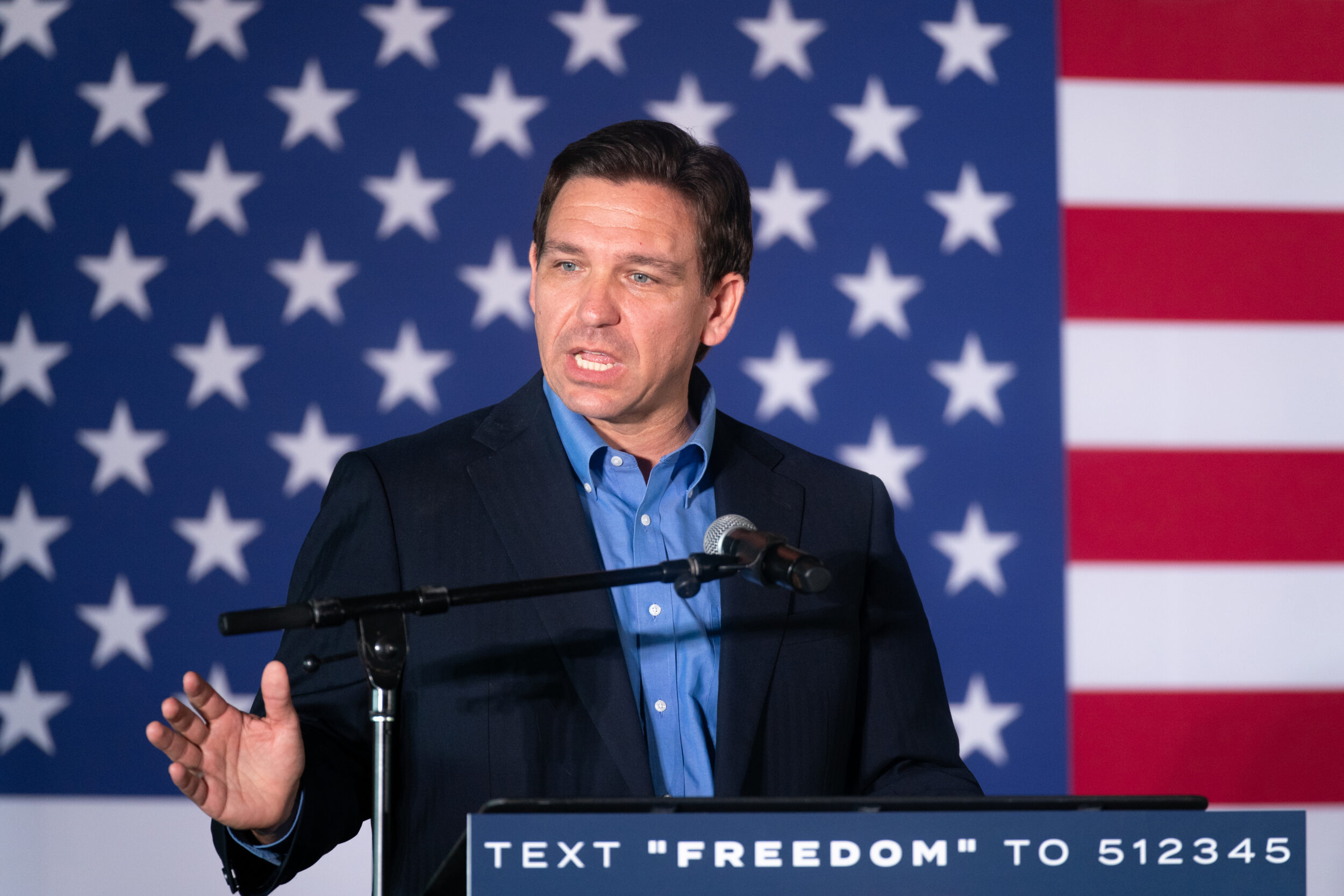Man in a blue shirt and blue suit stands with a microphone in front of the American flag