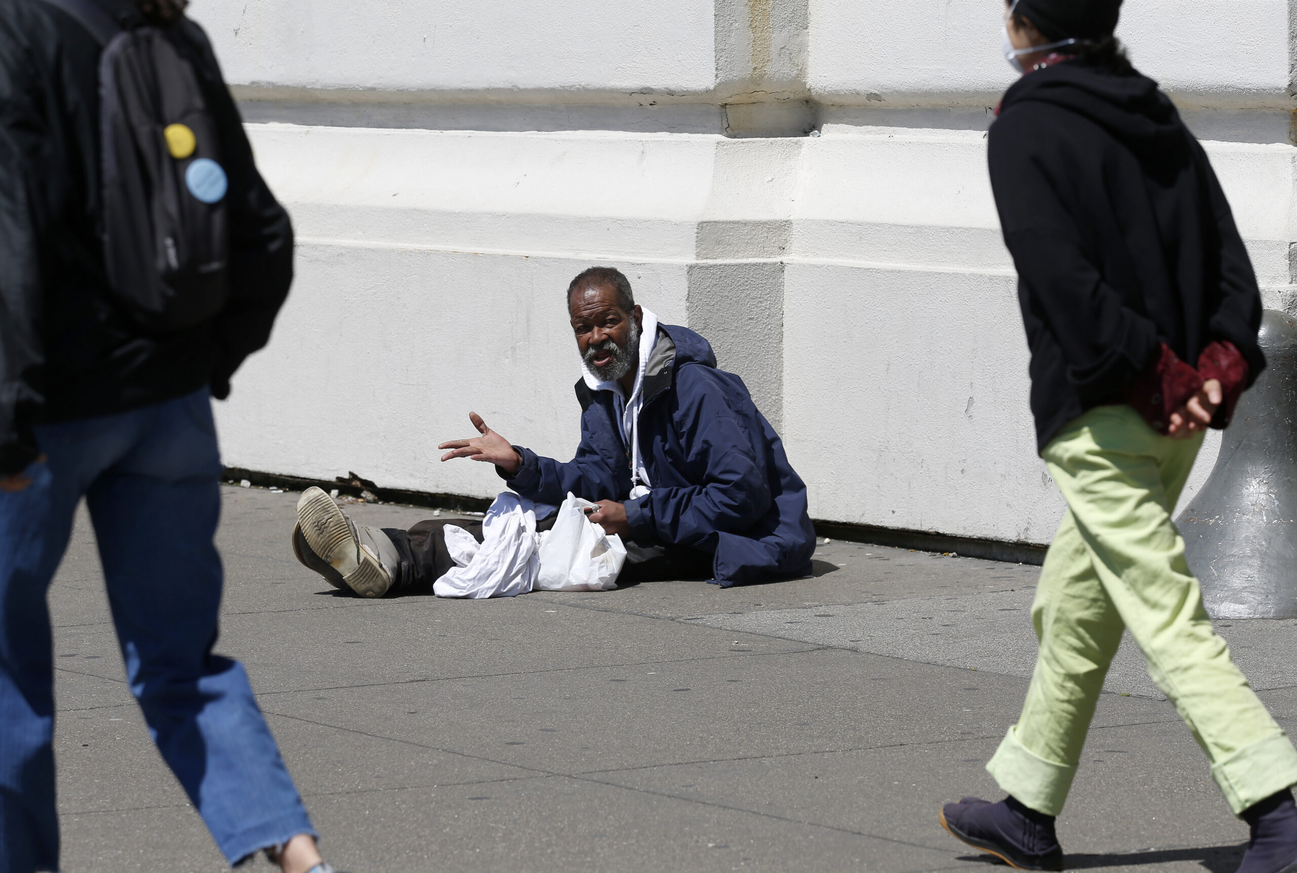 An unhoused person sits on sidewalk as others walk by,