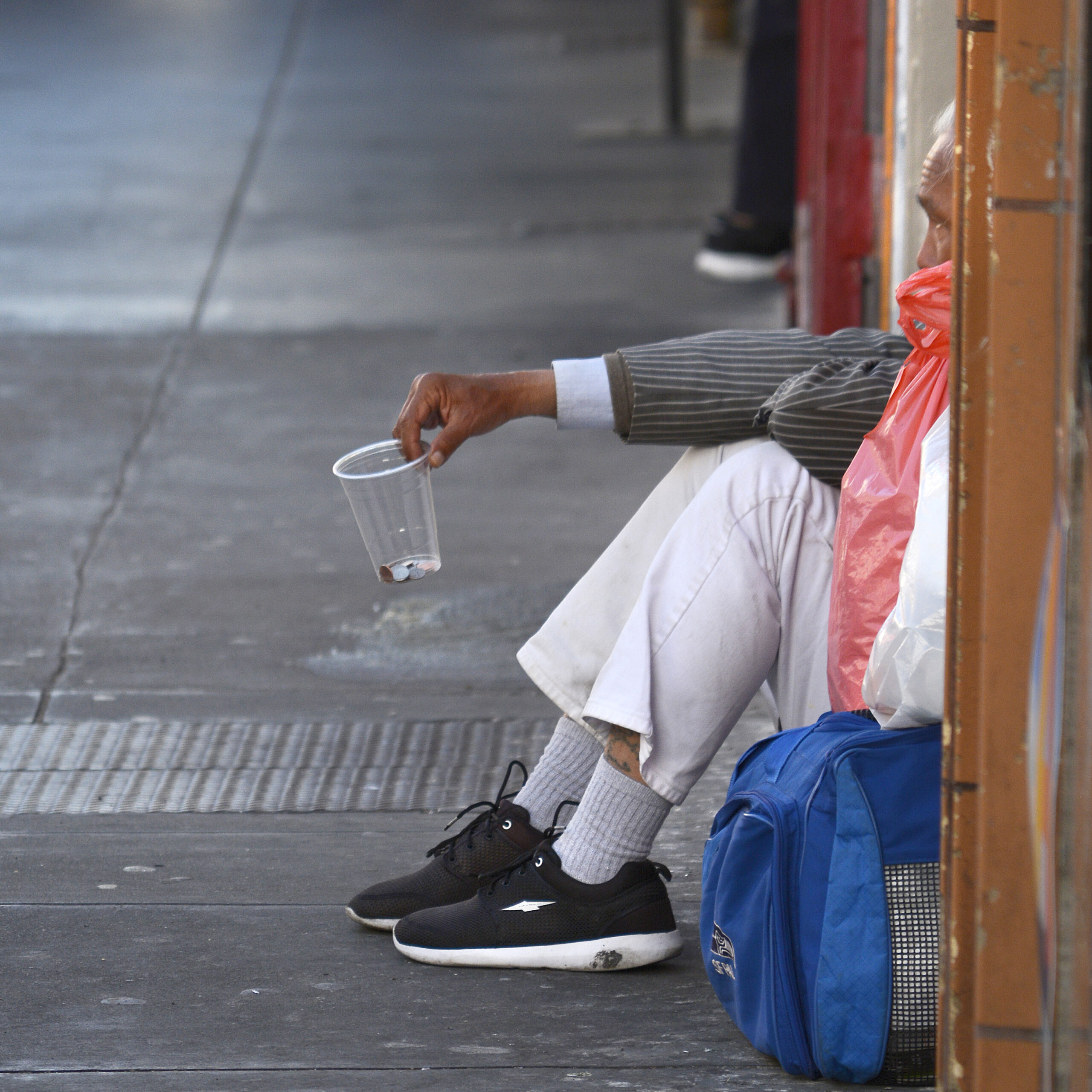 A person sitting on the sidewalk, holding out a cup with a few coins, beside a blue backpack.