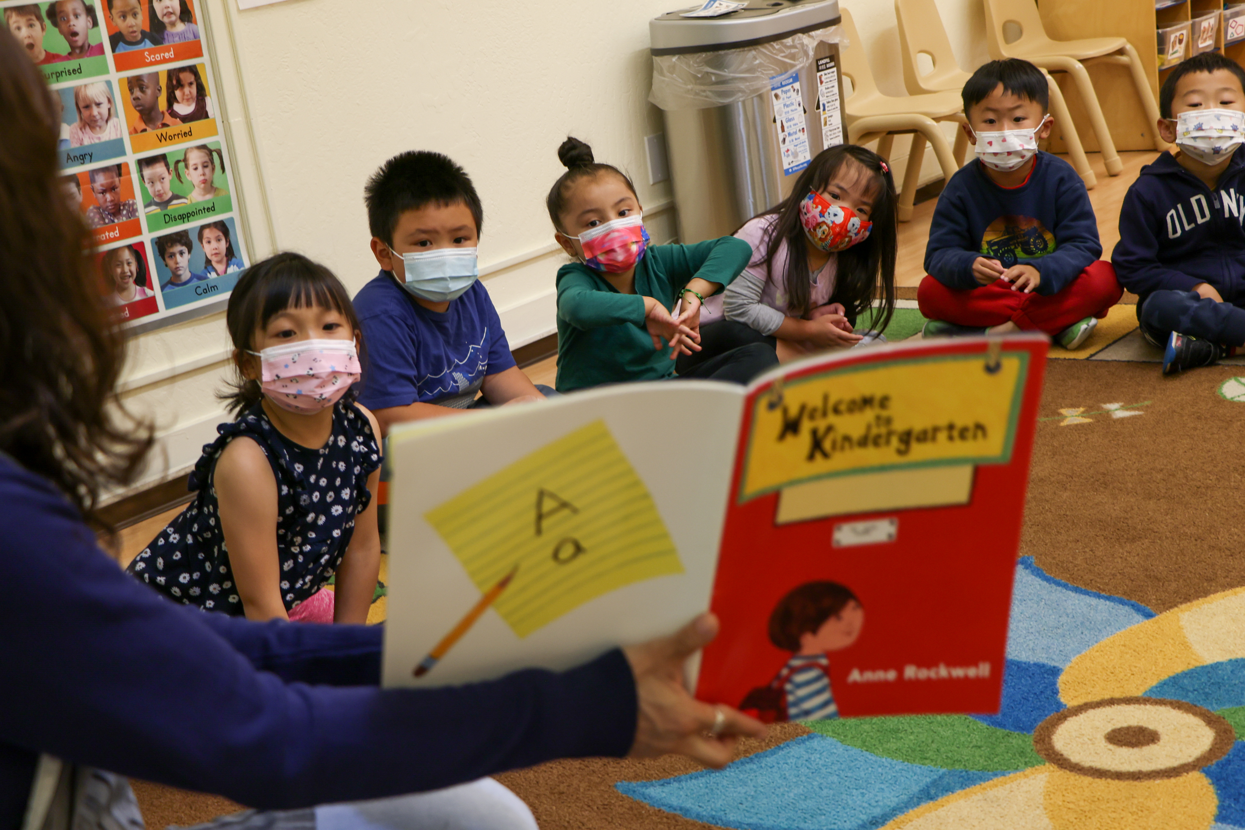 A teacher reads a "Welcome to Kindergarten" book to a group of young children sitting on a colorful carpet. The children wear face masks and listen attentively.