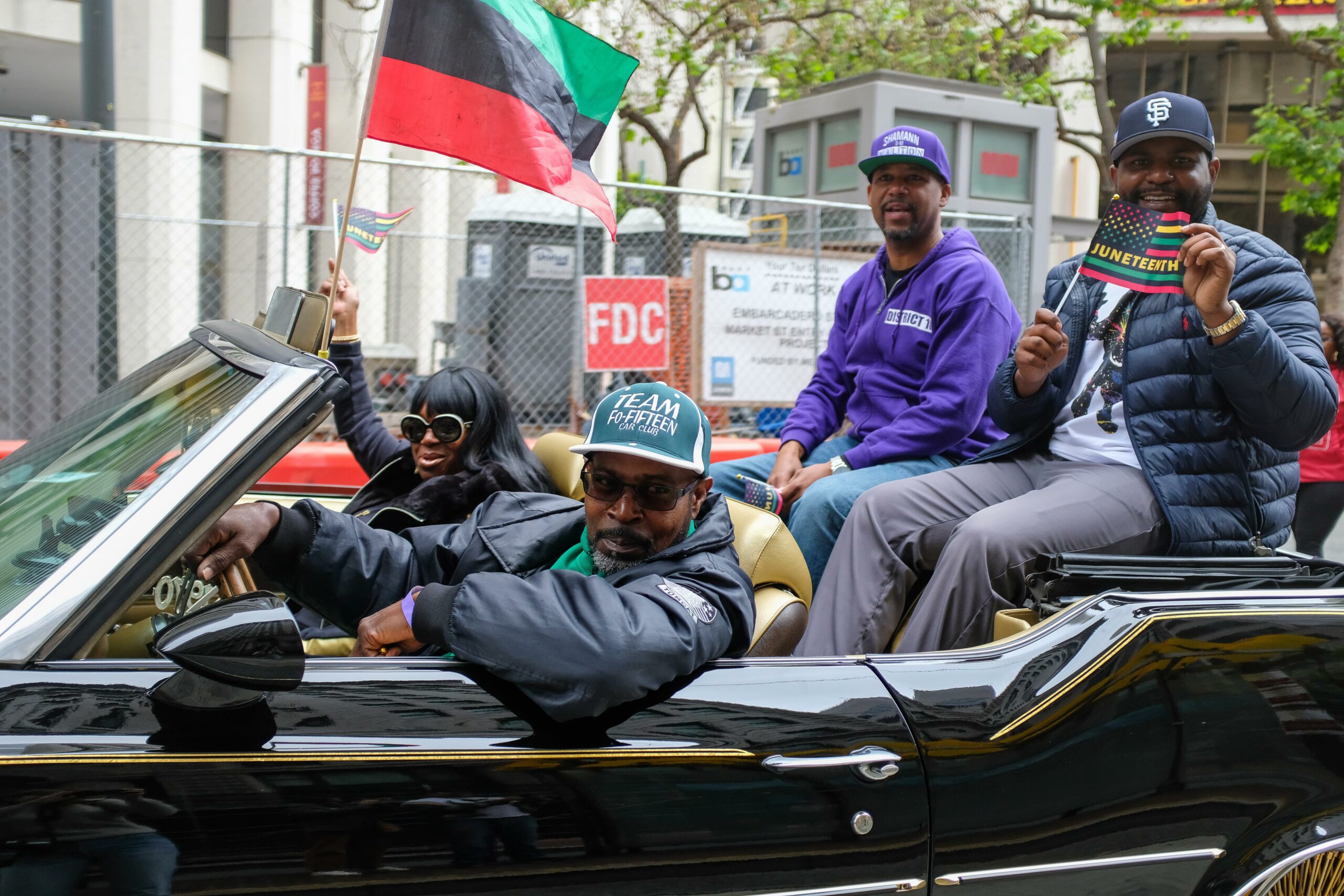 Four people are smiling in a black convertible car, holding a Pan-African flag and Juneteenth flag.