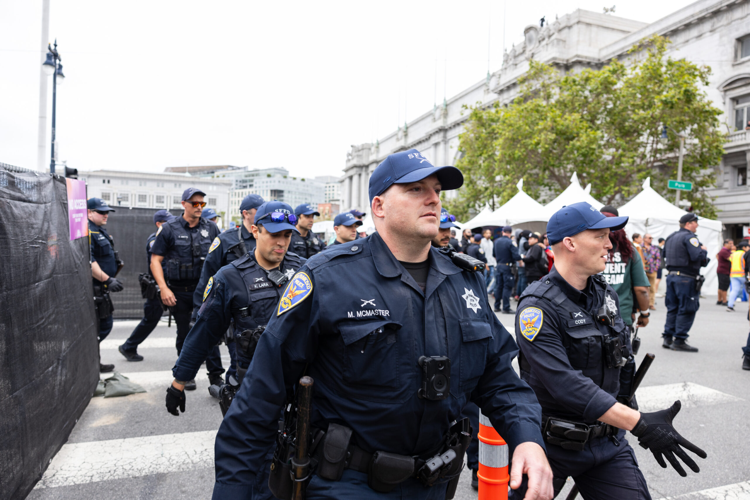 A group of police officers in uniform patrol an event, with a crowd and tents in the background.
