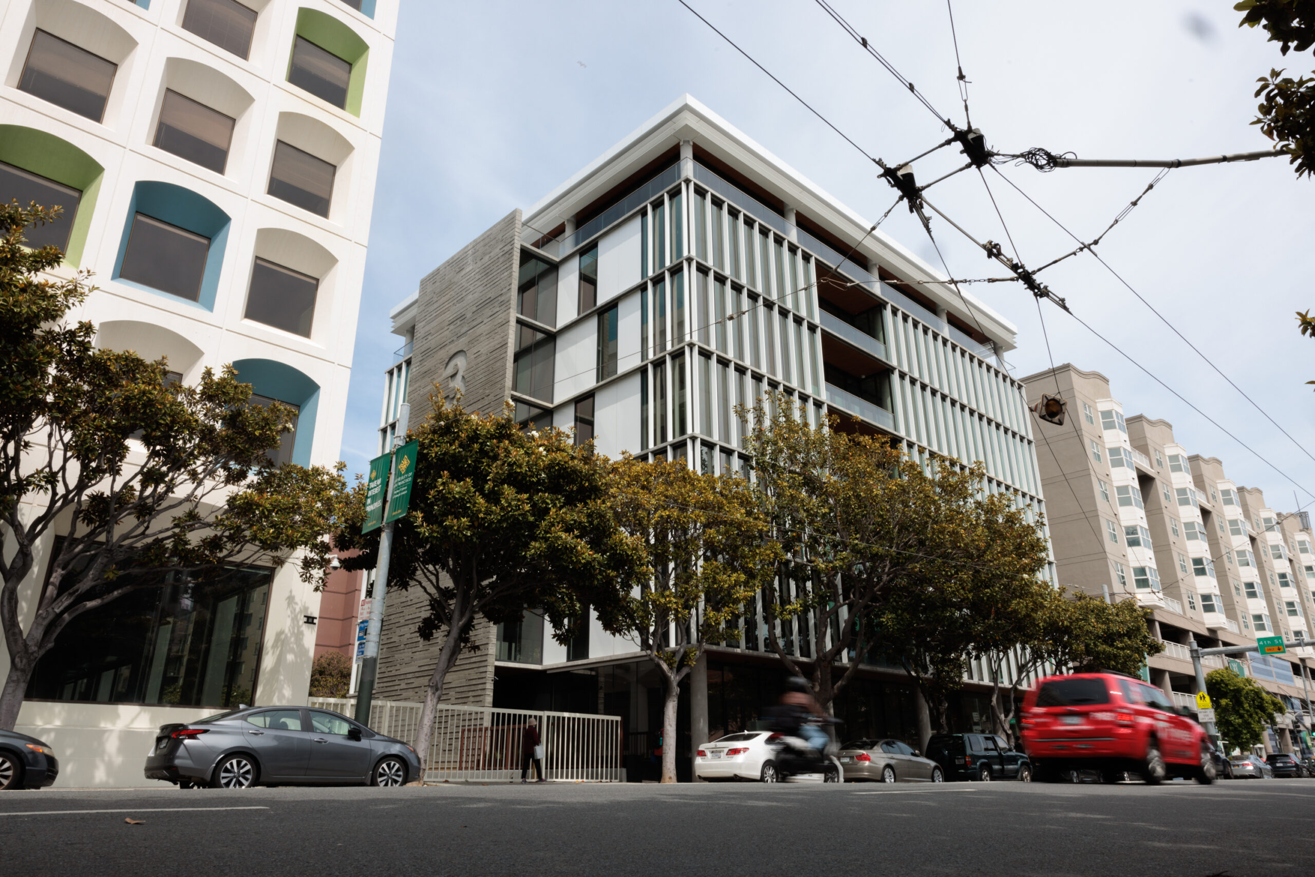 The image shows a modern urban street with tall, glassy buildings, trees lining the sidewalk, and cars, including a red SUV, moving on the road.