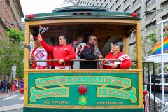 People in festive attire on a decorated cable car with &quot;VAN NESS AVE, CALIFORNIA &amp; MARKET STREETS&quot; signage.