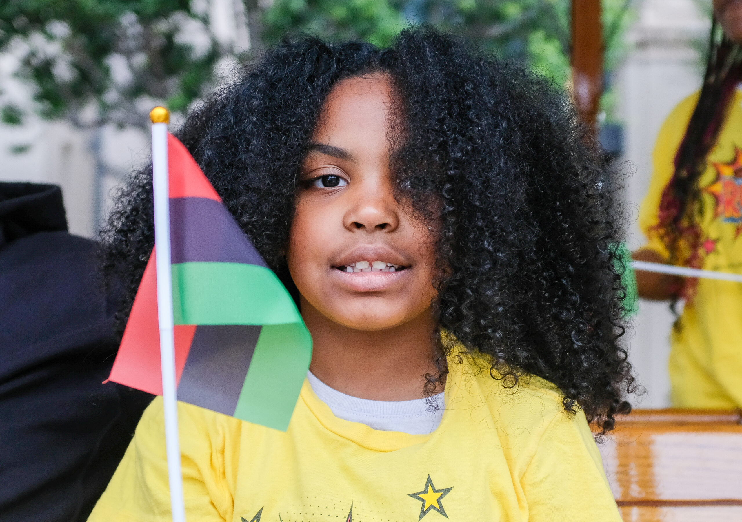 A child with curly hair holding a flag, wearing a yellow shirt with a star, smiles gently.