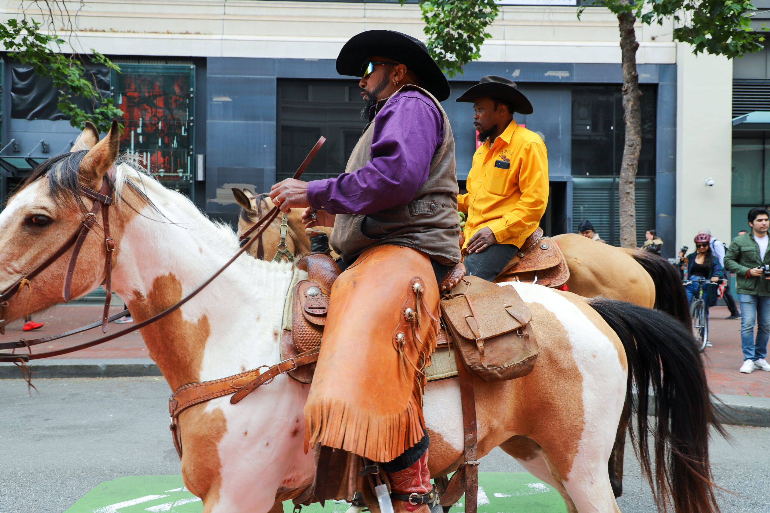 Two men in cowboy attire riding horses on a city street.