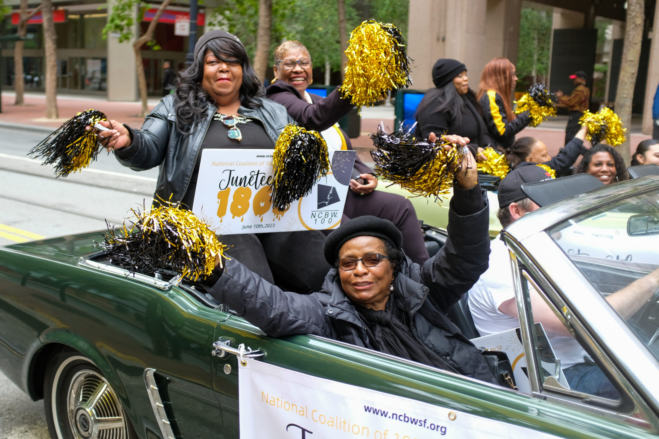 People in a car parade, holding pom-poms and a Juneteenth sign, celebrating with joyous expressions.
