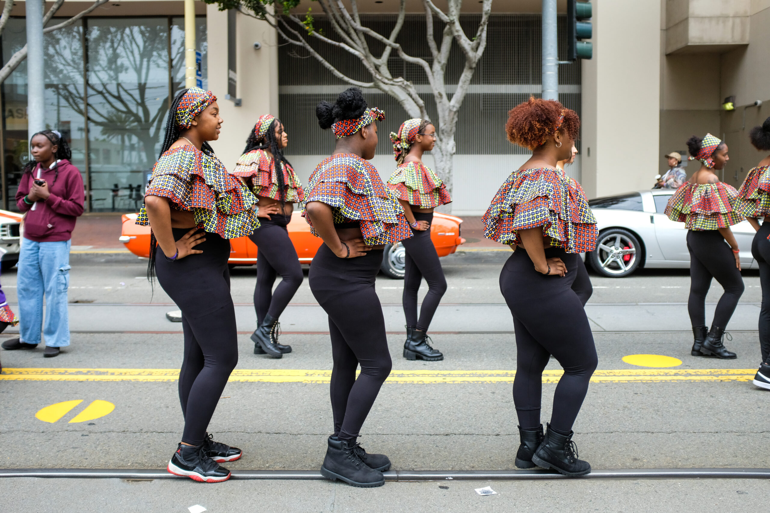 A group of dancers in matching tops and headwraps, black leggings, and boots are lined up on a street.