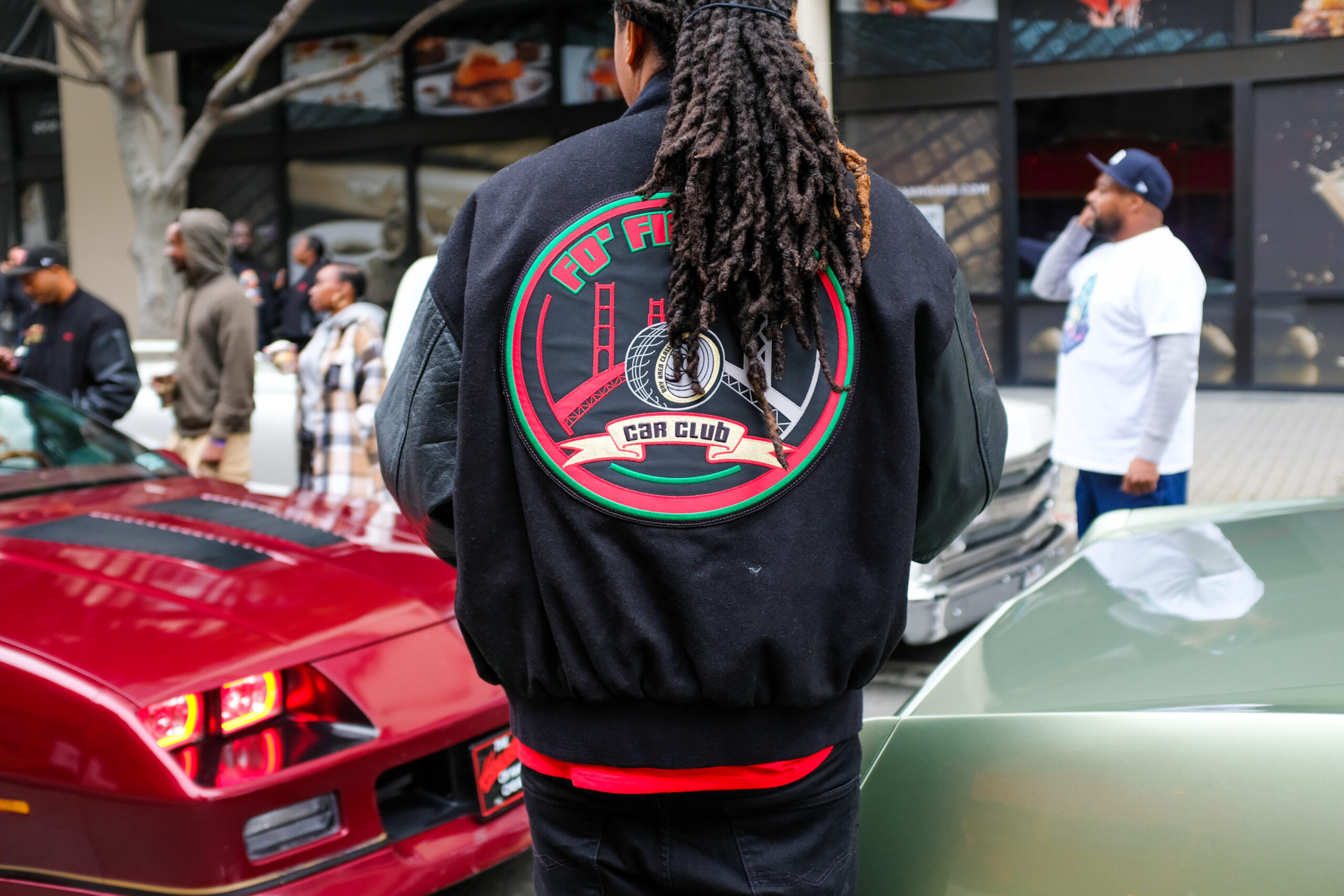 A person with dreadlocks is facing away, wearing a jacket with a car club emblem, in front of two vintage cars and onlookers.