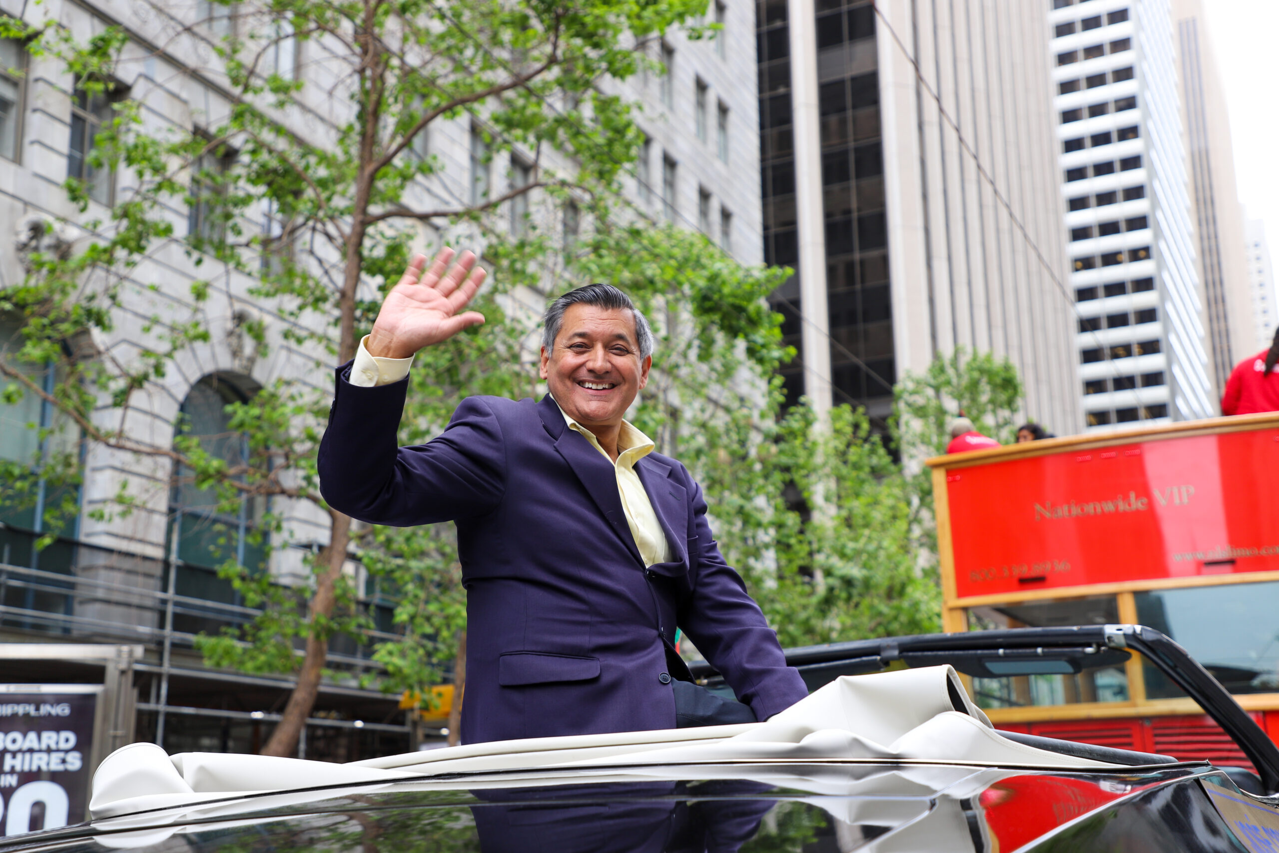 A person in a suit waving from a convertible, with trees and high-rise buildings in the background.
