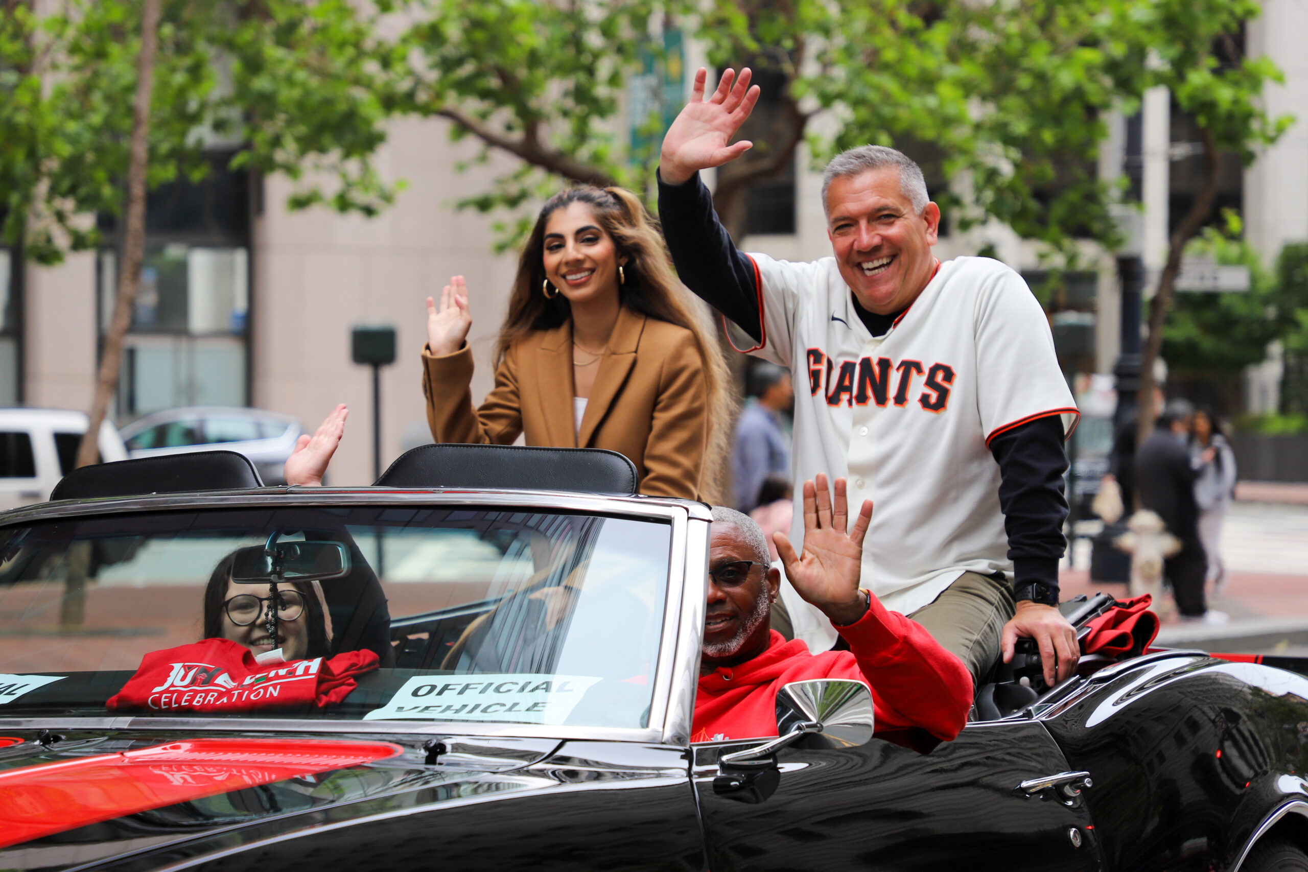Three people are joyfully waving from a convertible vehicle, with one wearing a Giants jersey.