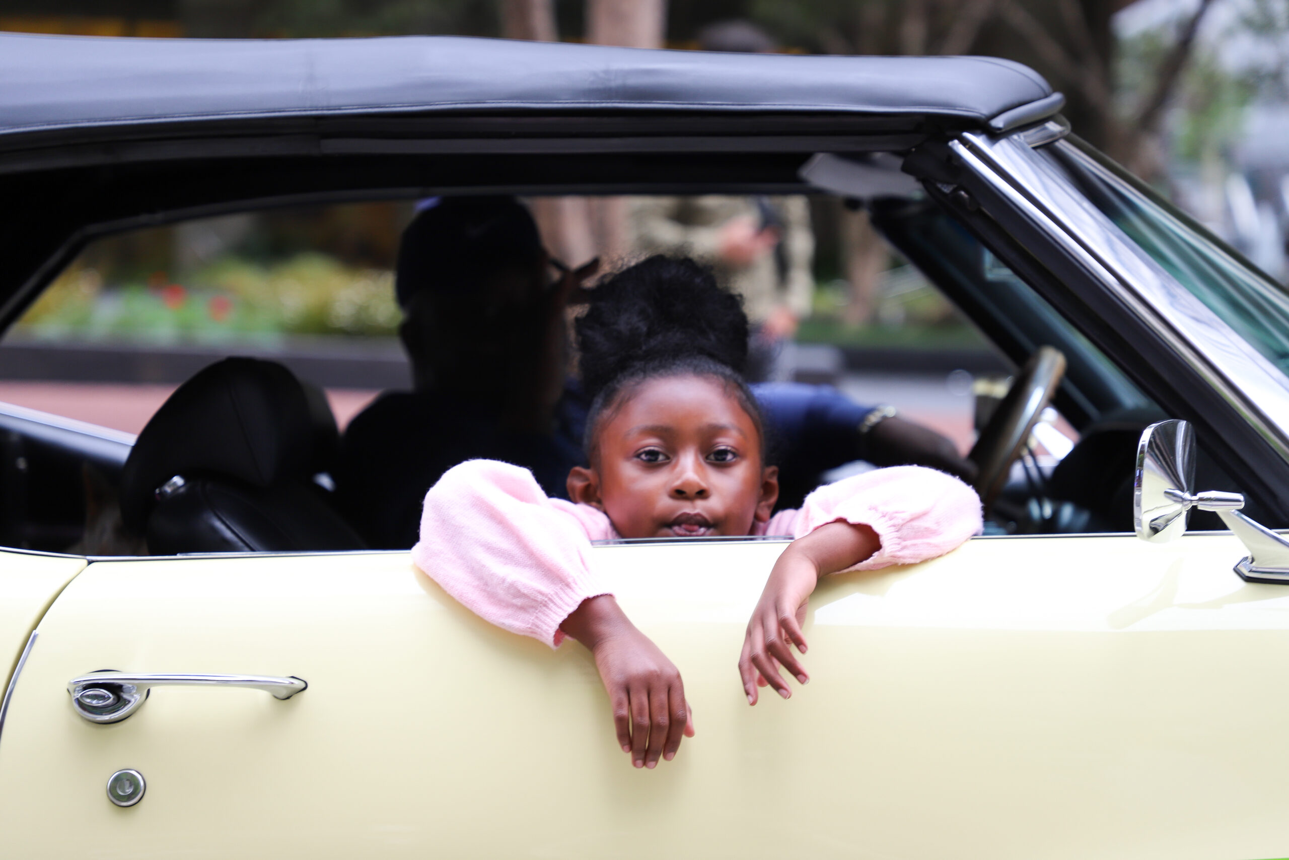 A young girl leans on the door of a classic yellow convertible car, looking at the camera.