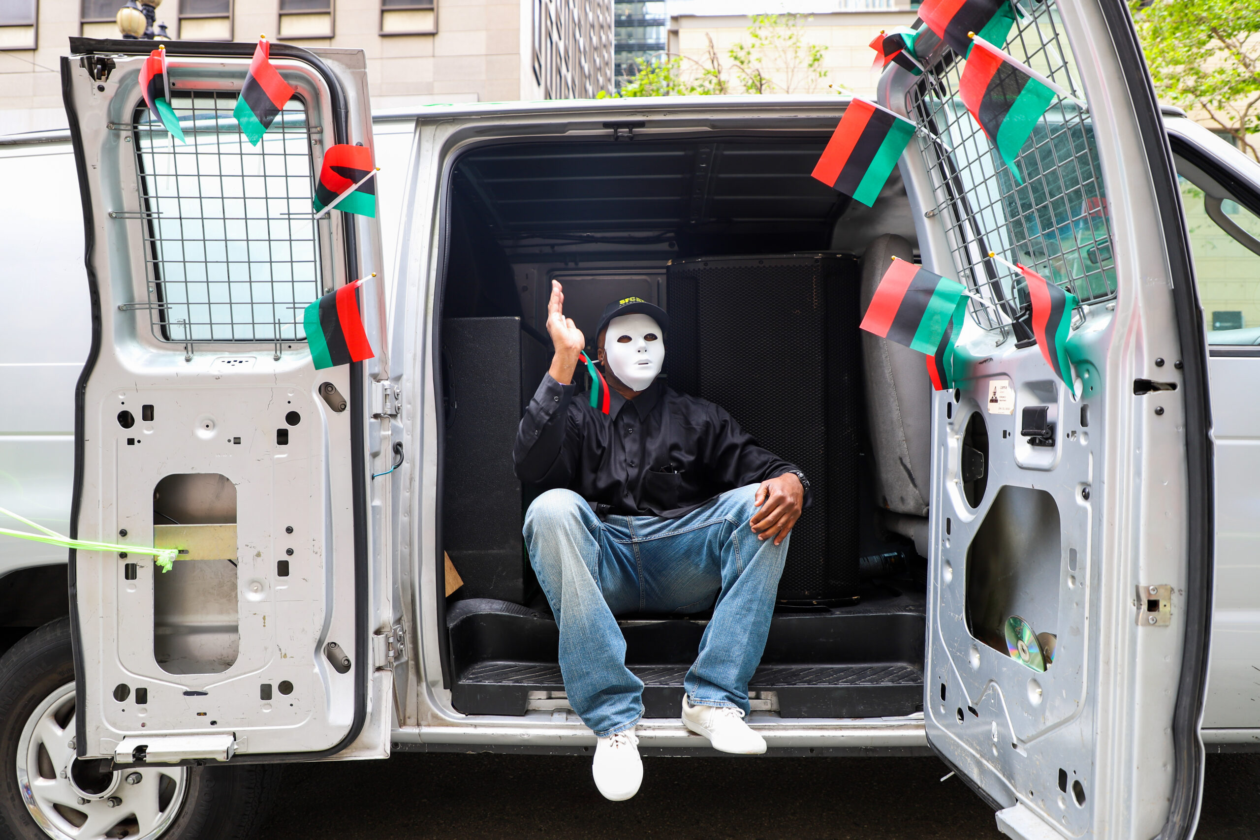 A person in a white mask sits in an open van door, gesturing, with red, black, and green flags attached around them.