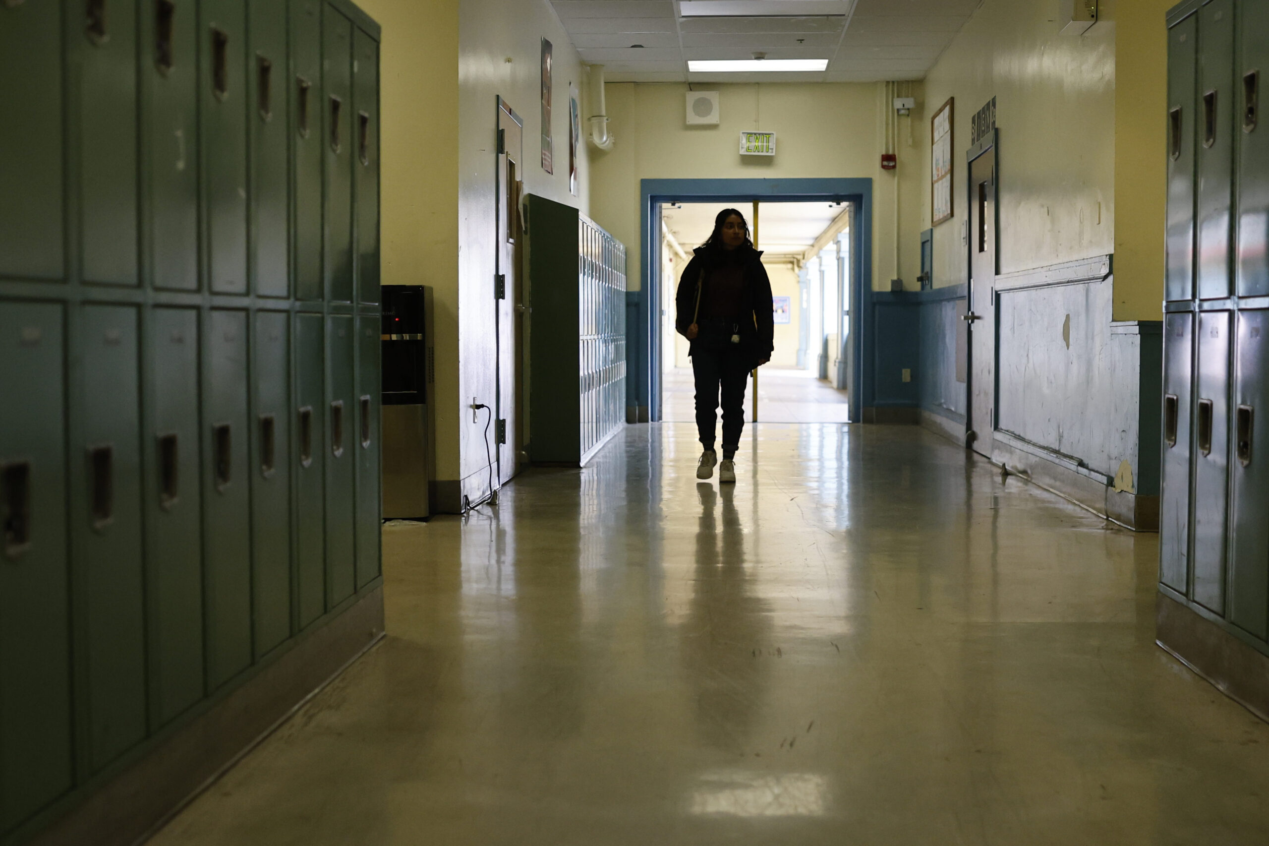 A person walks down a dimly lit school hallway lined with green lockers on both sides, with a door at the end leading to a brighter area.