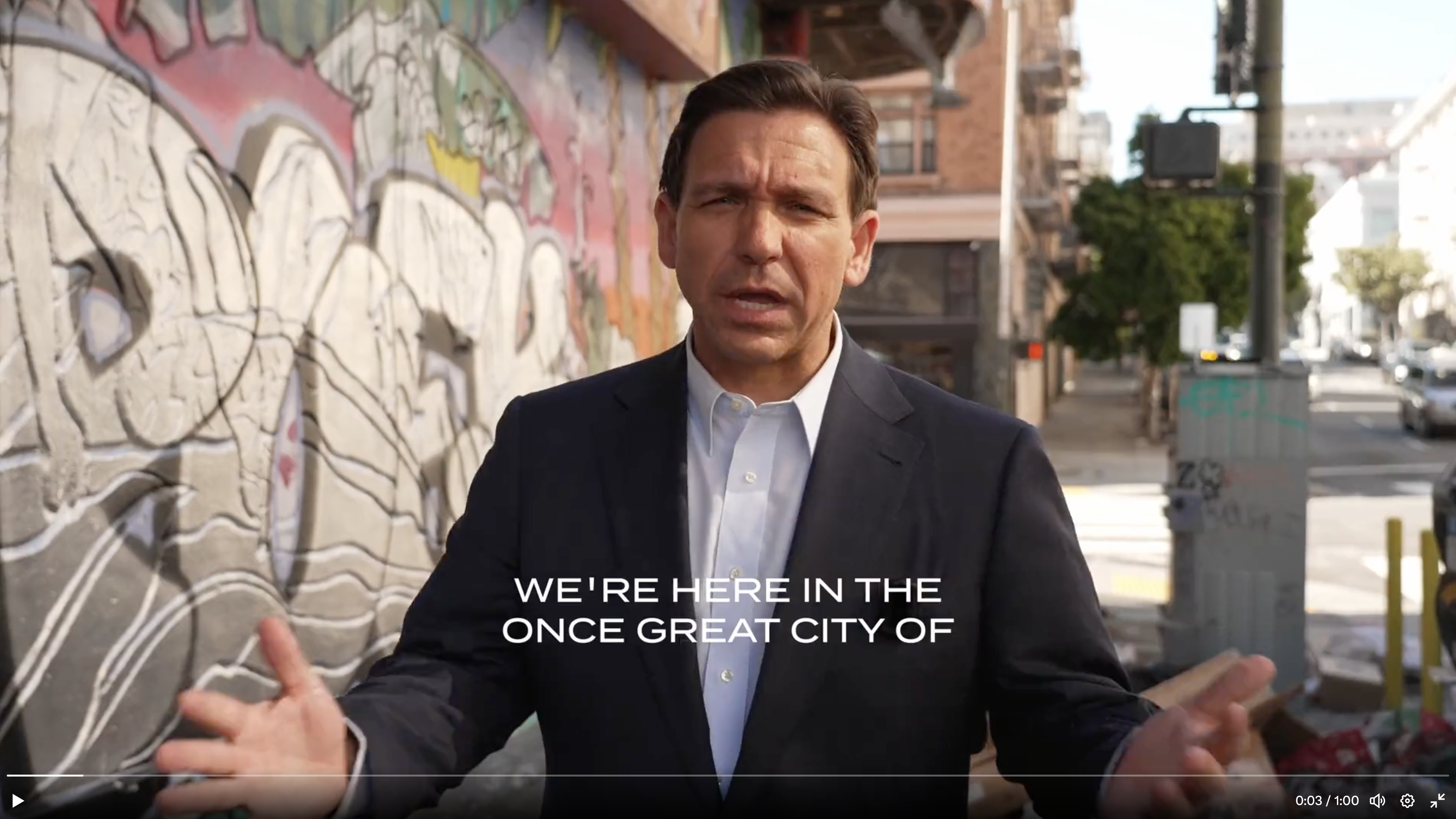 A screenshot of a Ron DeSantis campaign ad shows the Florida governor in a white button-down shirt and black suit jacket with no tie, gesturing with his arms while he stands next to a graffiti-covered building in San Francisco.