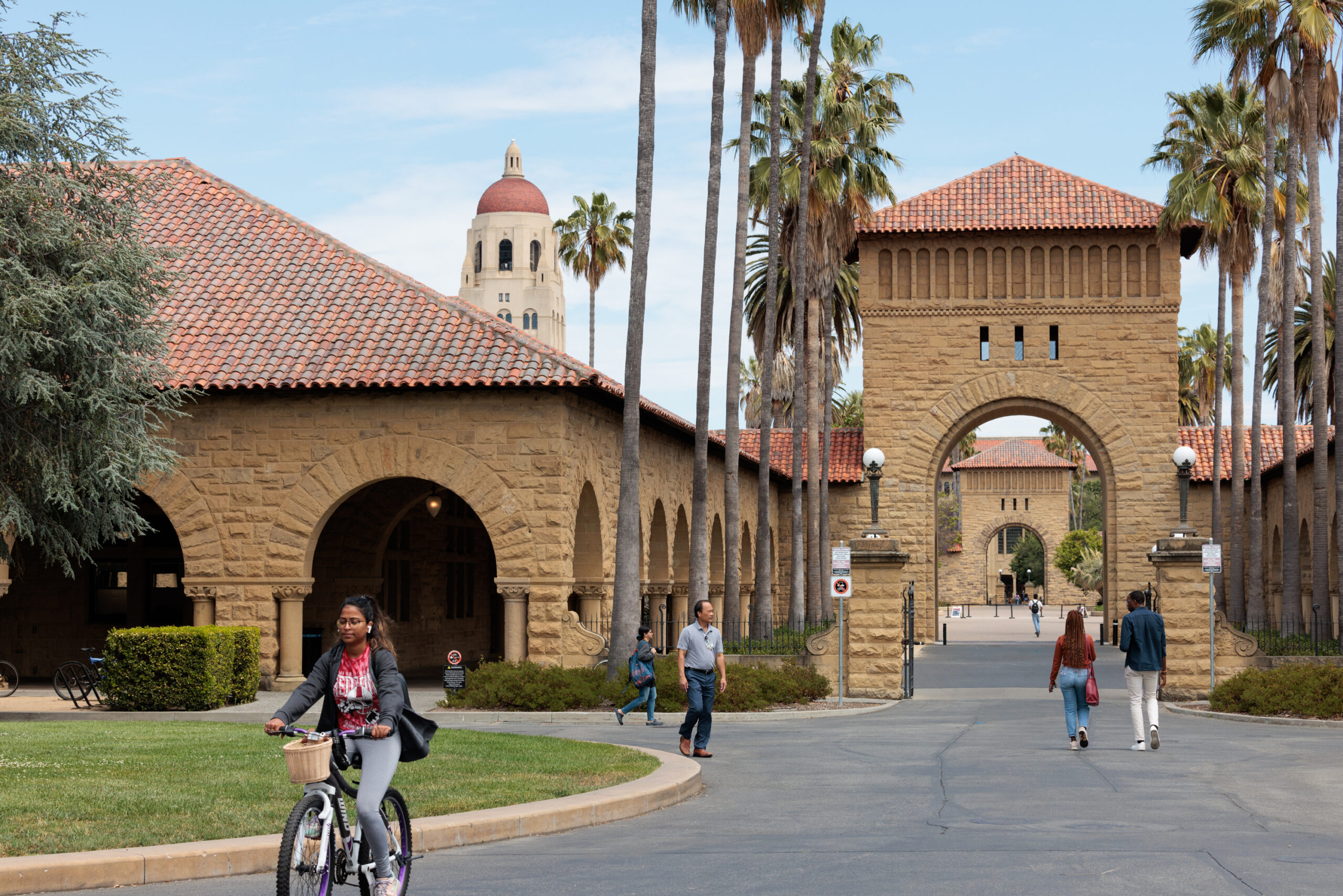 A woman rides a bicycle near Spanish-style architecture, with an arched walkway and a tall tower in the background, surrounded by palm trees.