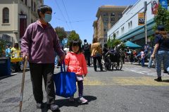 An elderly person holding hands with a child on a sunny street among other pedestrians.