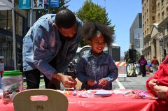 A man and a smiling young girl are at a craft table on a sunny street, both in denim jackets.