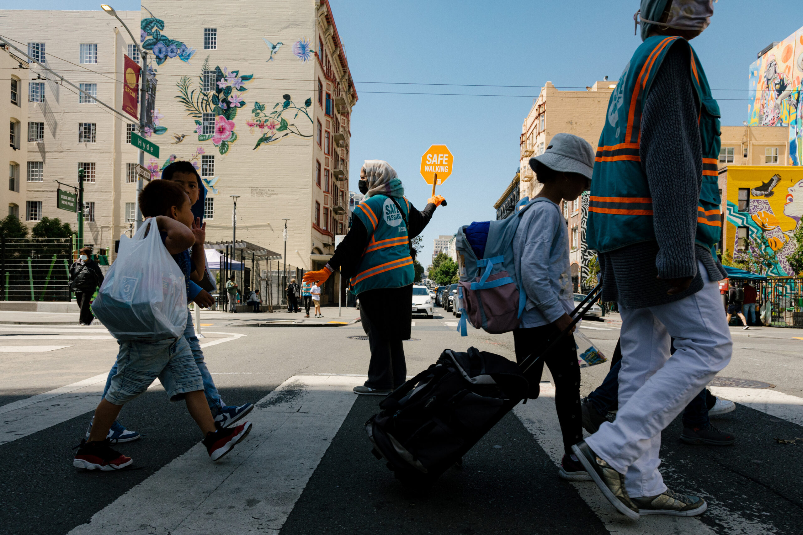 A crossing guard holds a &quot;SAFE WALKING&quot; sign as pedestrians cross a sunny urban street with vivid murals.