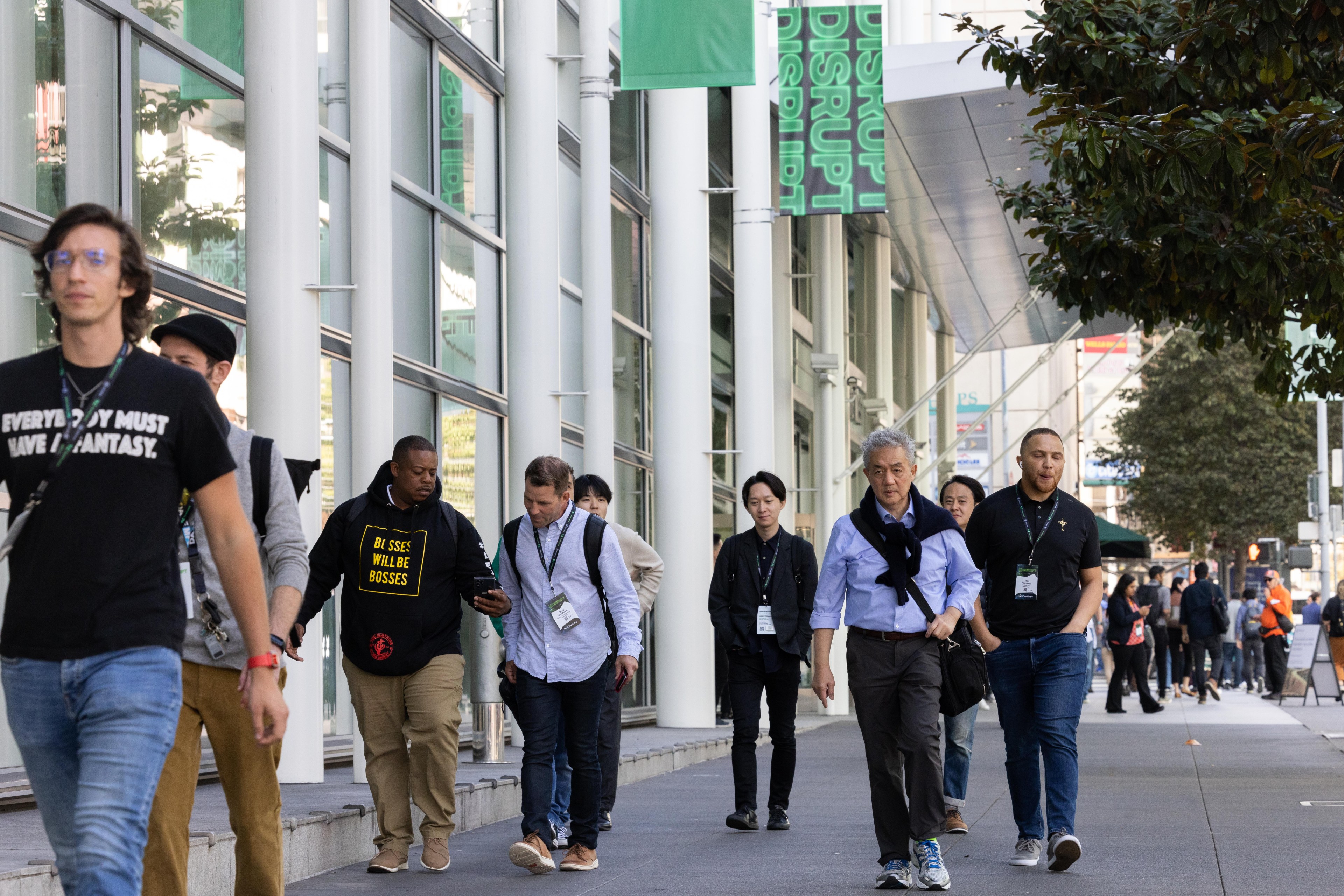 A group of people walk past a building with green &quot;DISRUPT&quot; banners, some wearing conference badges. The area is lively, with trees and large windows.