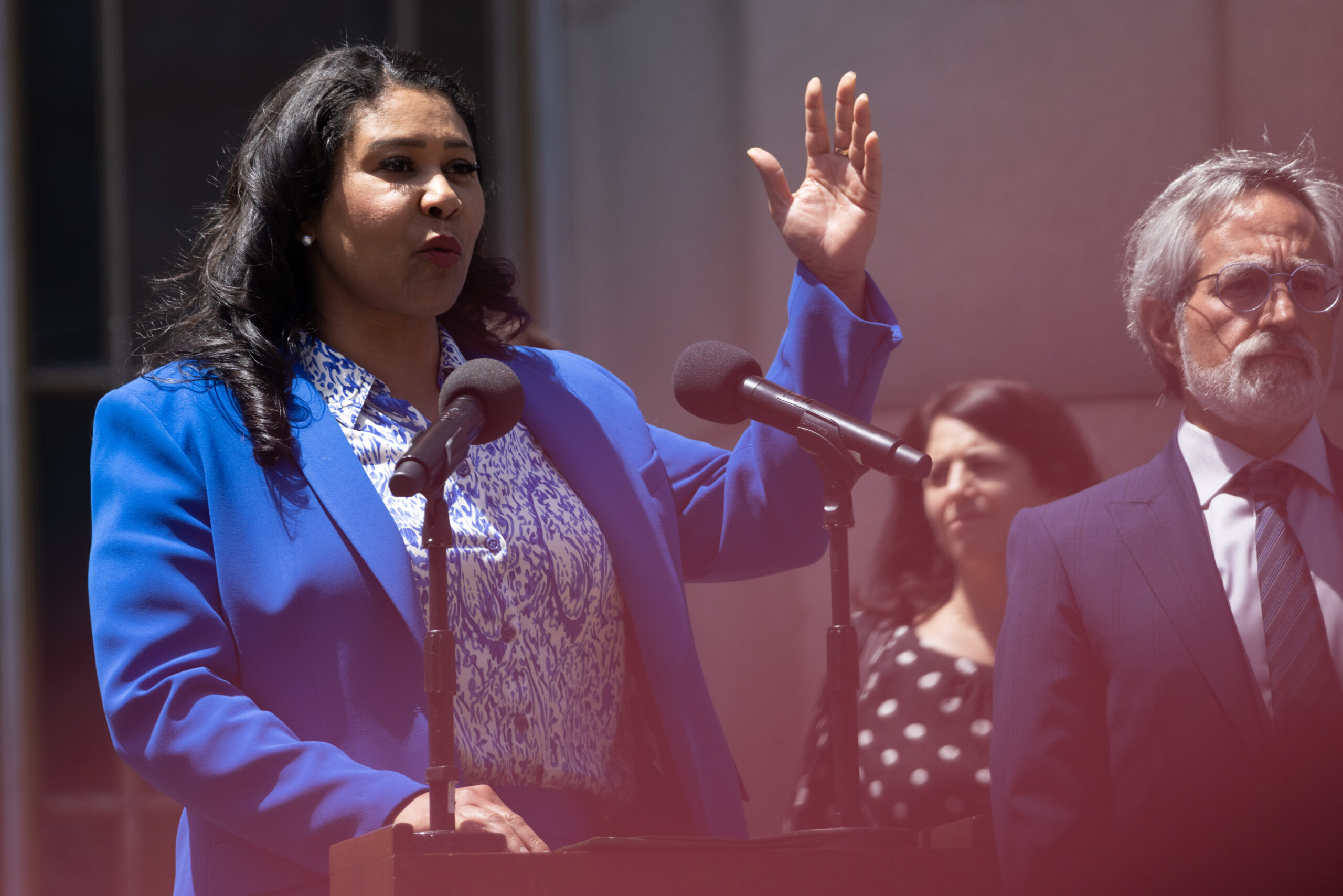 A woman in a blue blazer speaks at a podium, raising her hand, with two people behind her.