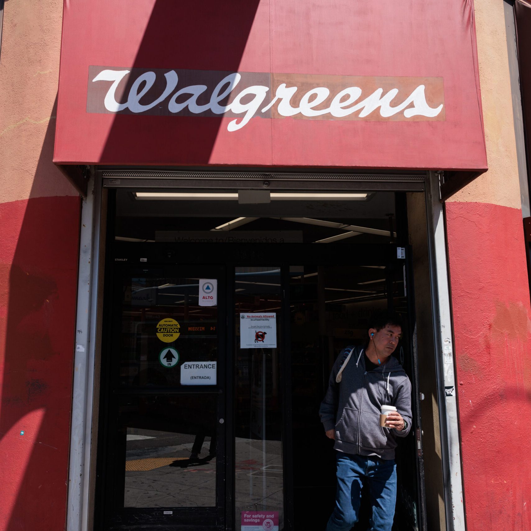 A person is exiting a Walgreens store, wearing a dark hoodie and carrying a drink. The storefront features a red awning with the Walgreens logo.